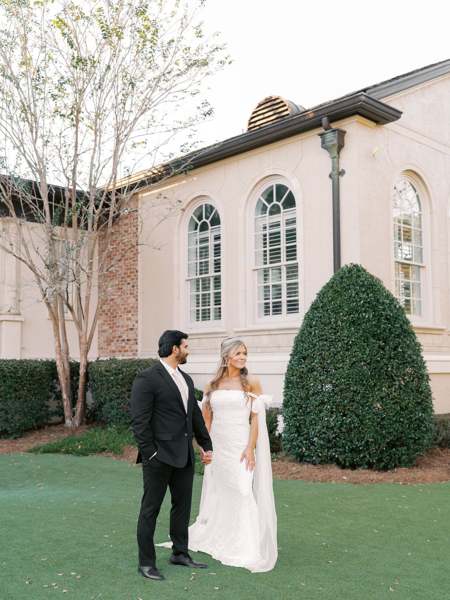 Wedding couple walks together on lawn beside traditional brick and white building.