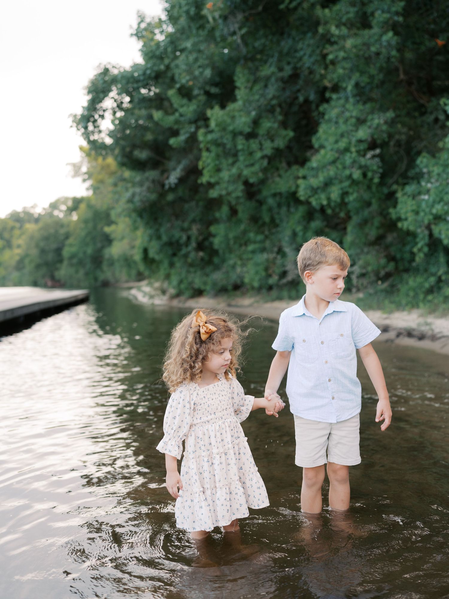 Two young children playing and splashing together in shallow water at the edge of a river on a sunny day.