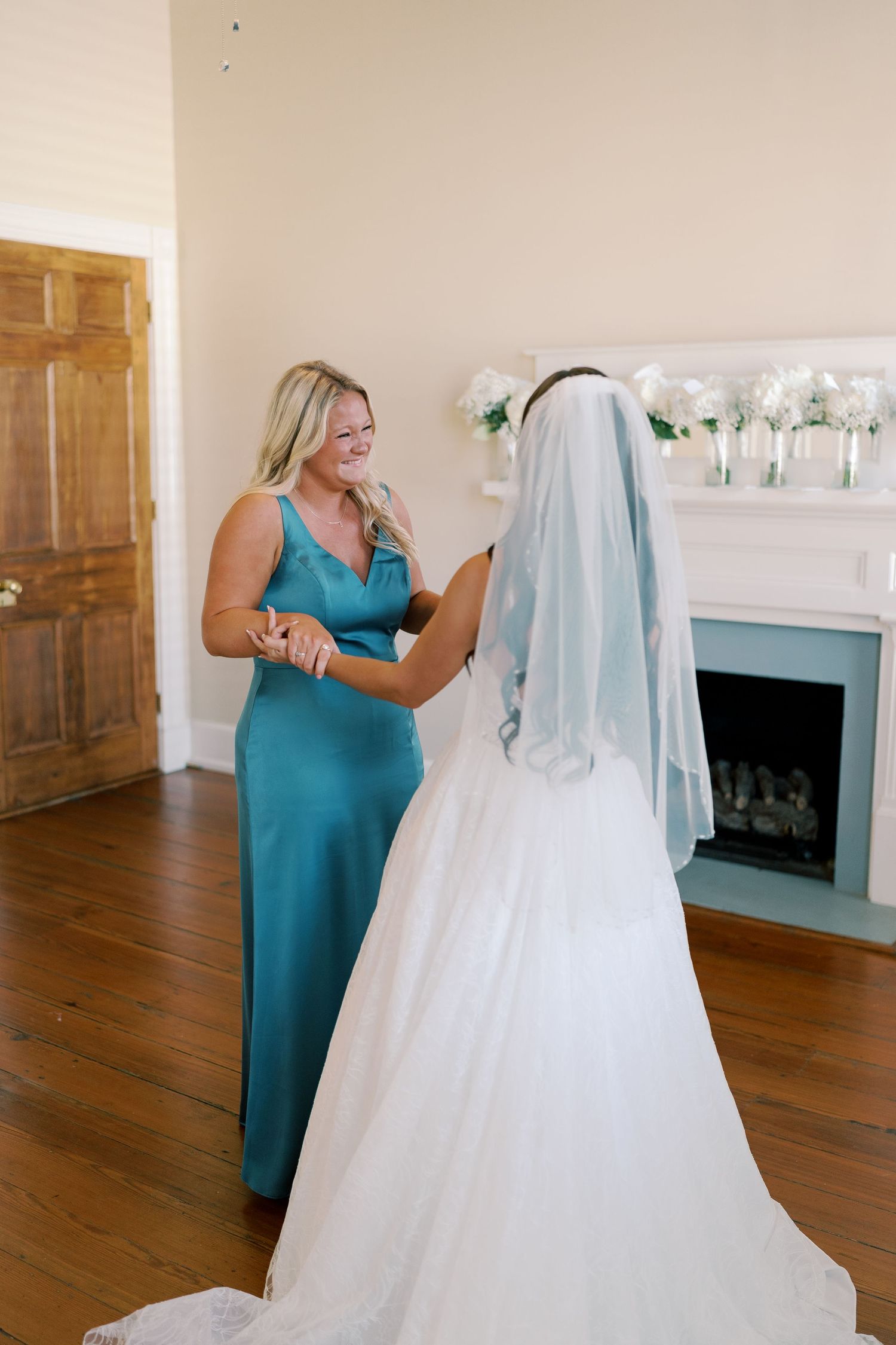 Bridesmaid in teal dress helps with wedding gown preparations in a room with hardwood floors.