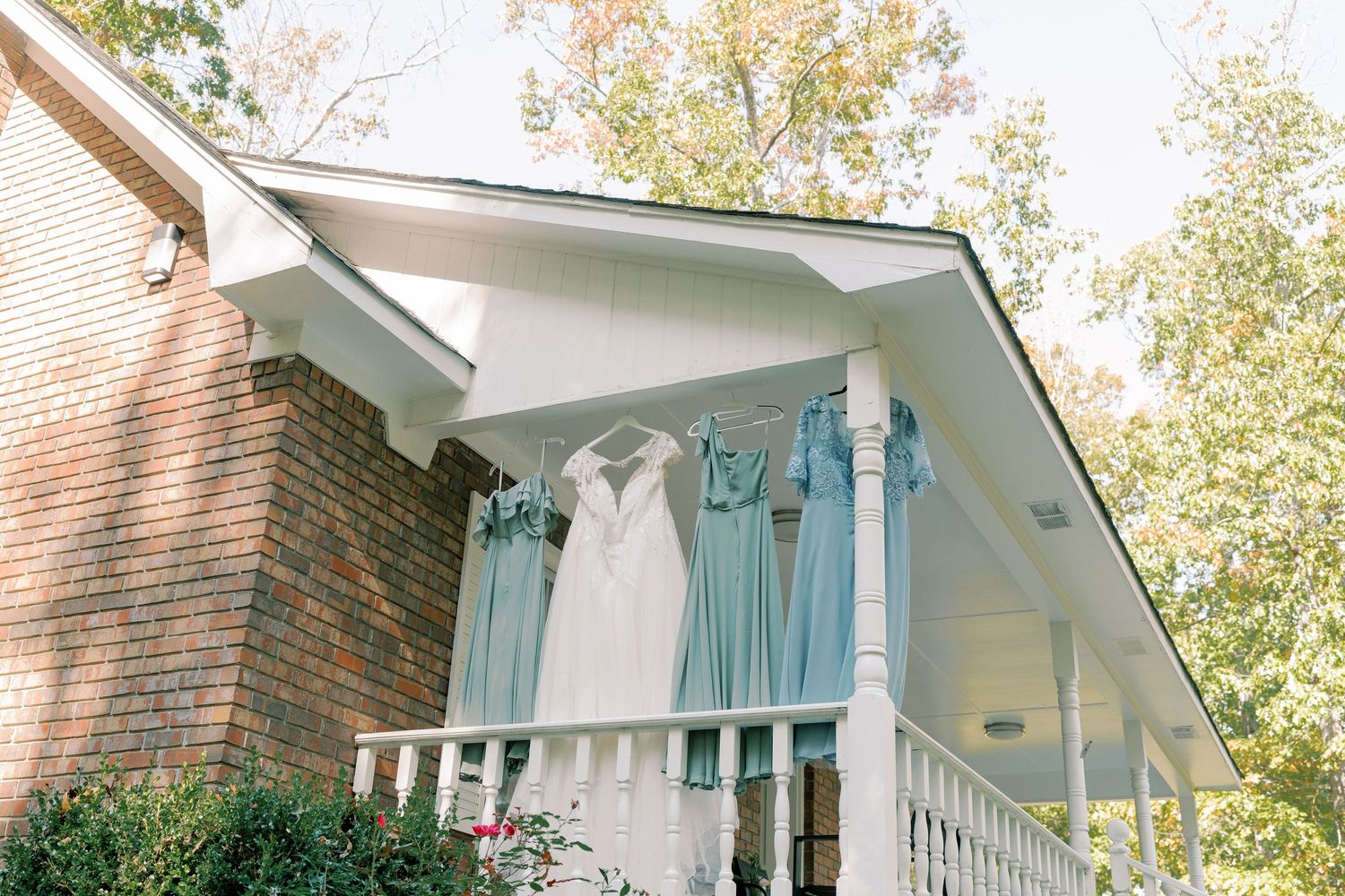 Wedding and bridesmaid dresses displayed on front porch of brick house with white railings.