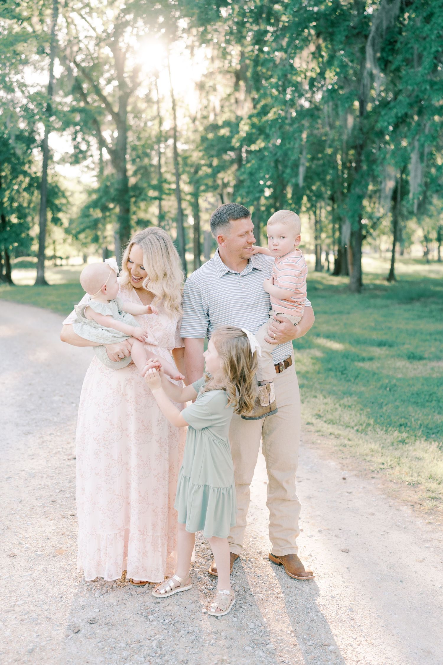 Family group photo session on a gravel path surrounded by green trees and golden sunlight.