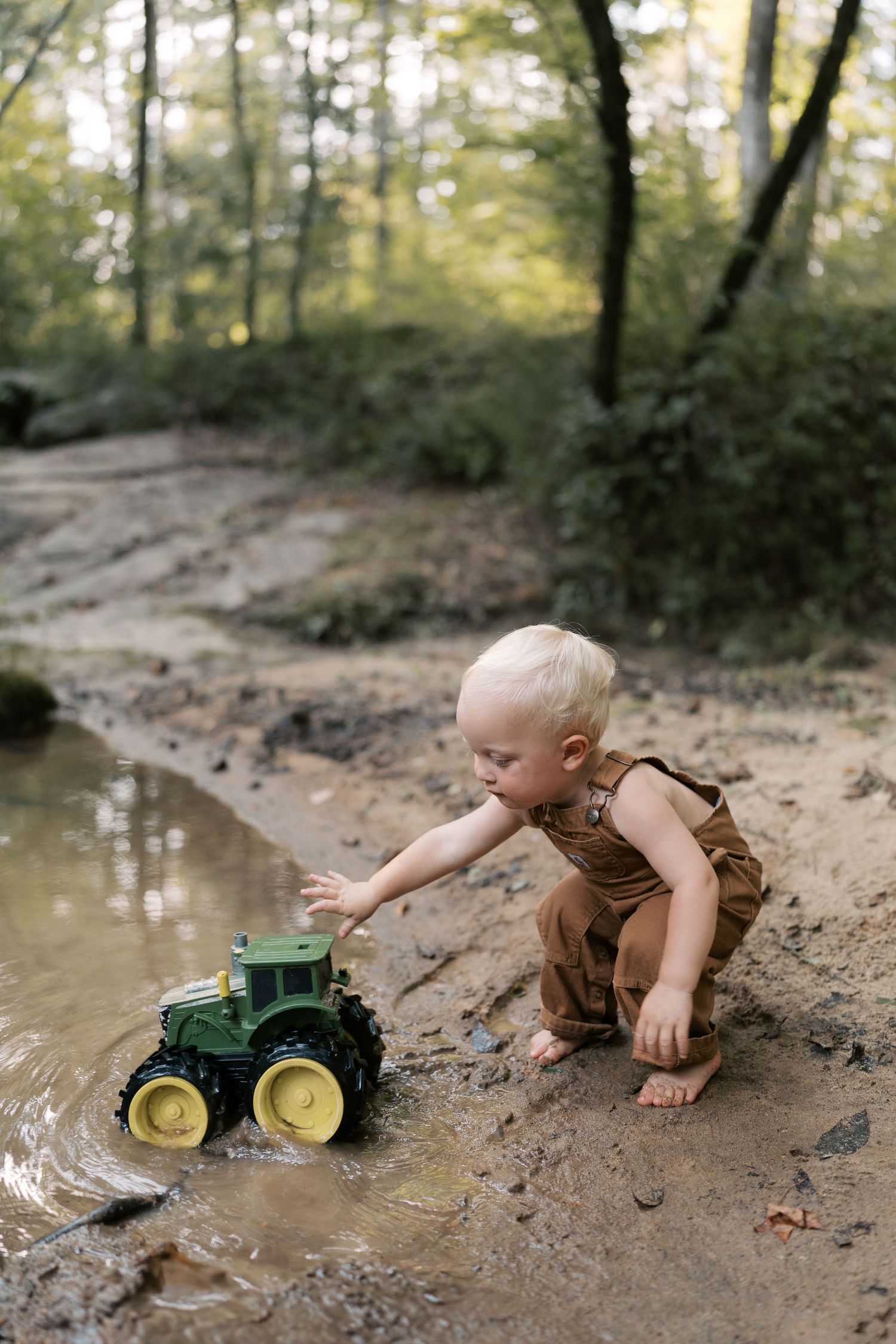 A toddler plays with a green toy tractor near a muddy creek bank in the woods.