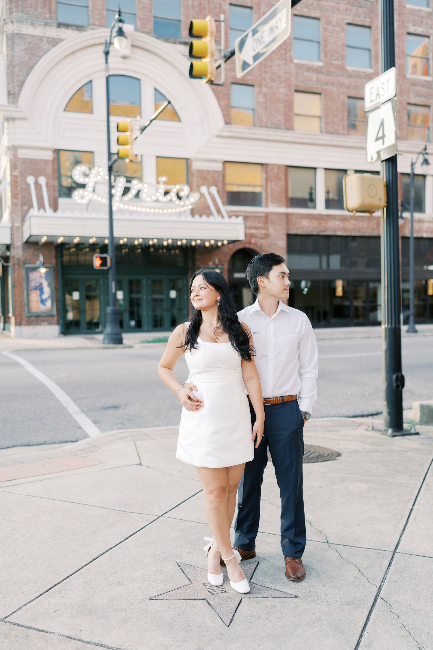 A couple shares a romantic moment on a city sidewalk with brick buildings and traffic lights in the background.