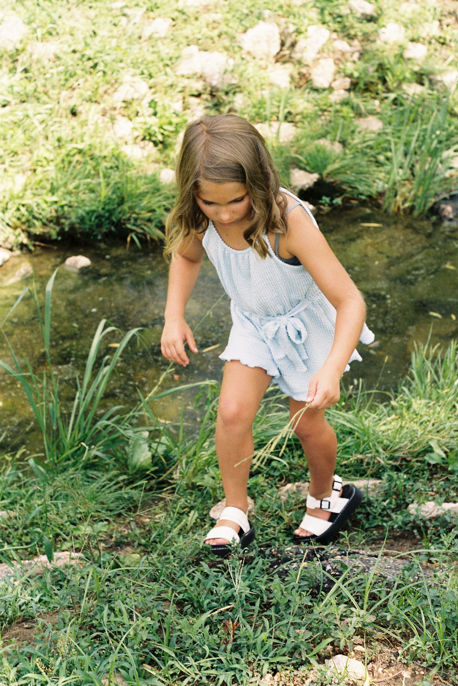 Young child in white dress exploring by a stream on a sunny day.