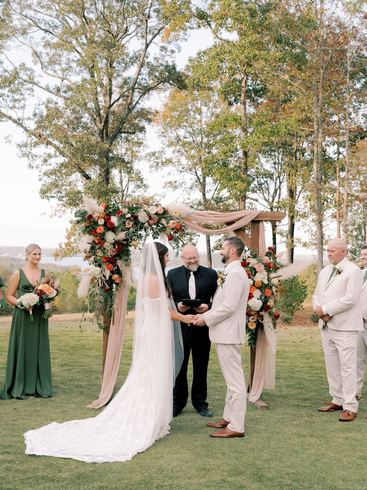 Wedding ceremony in progress with bridal party in green dresses and beige suits on a lawn.