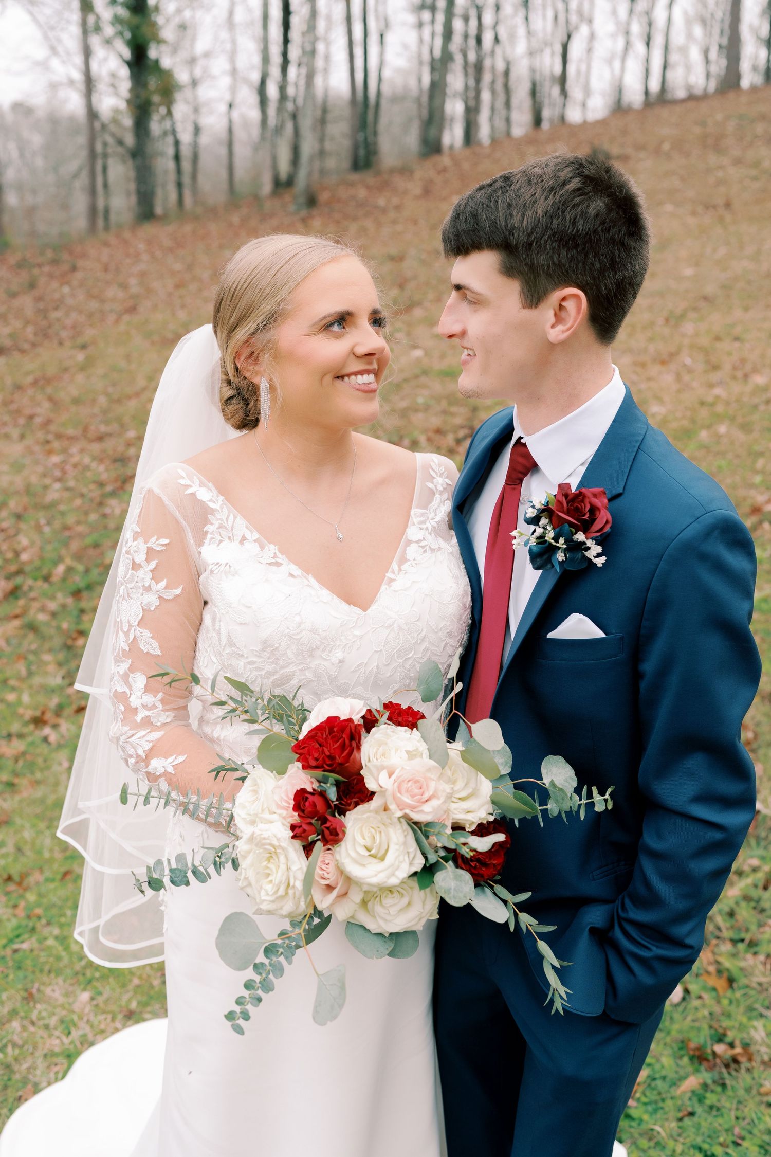Newlyweds pose with burgundy and cream bouquet against autumn landscape.