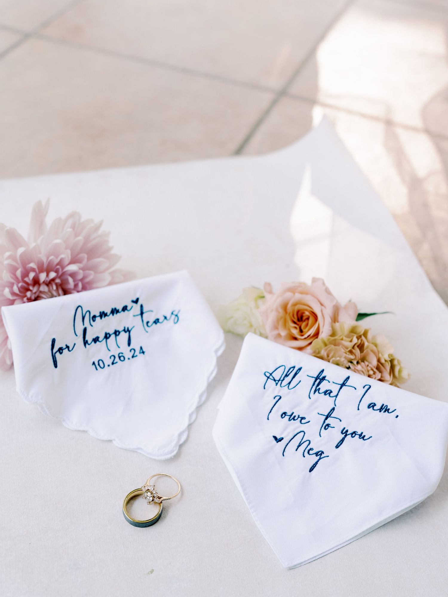 Handwritten blue calligraphy notes and wedding rings on white paper with soft pink flowers.