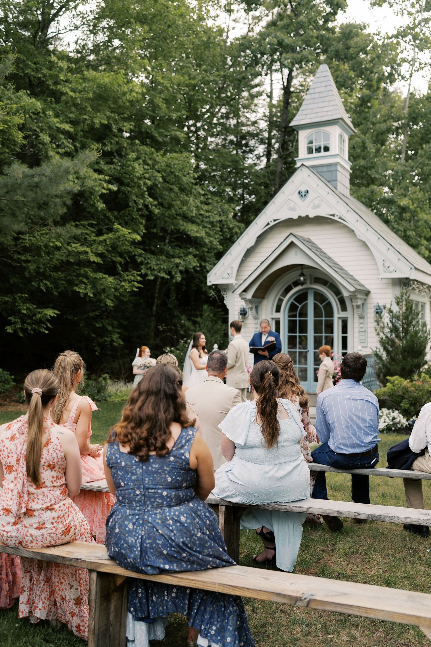 Guests watch a wedding ceremony from wooden benches outside a picturesque white chapel.
