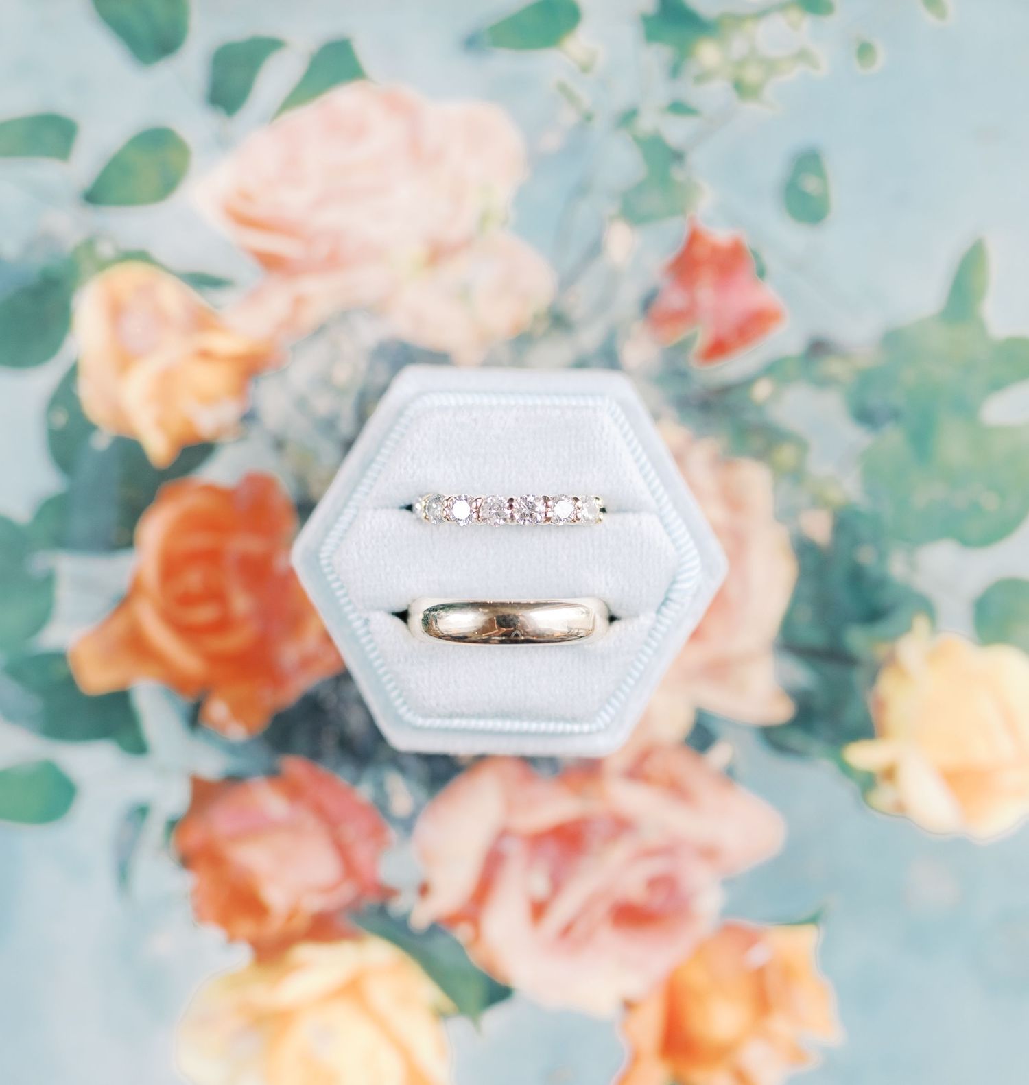 Wedding rings displayed on a white textured surface with soft peach and mint floral backdrop.