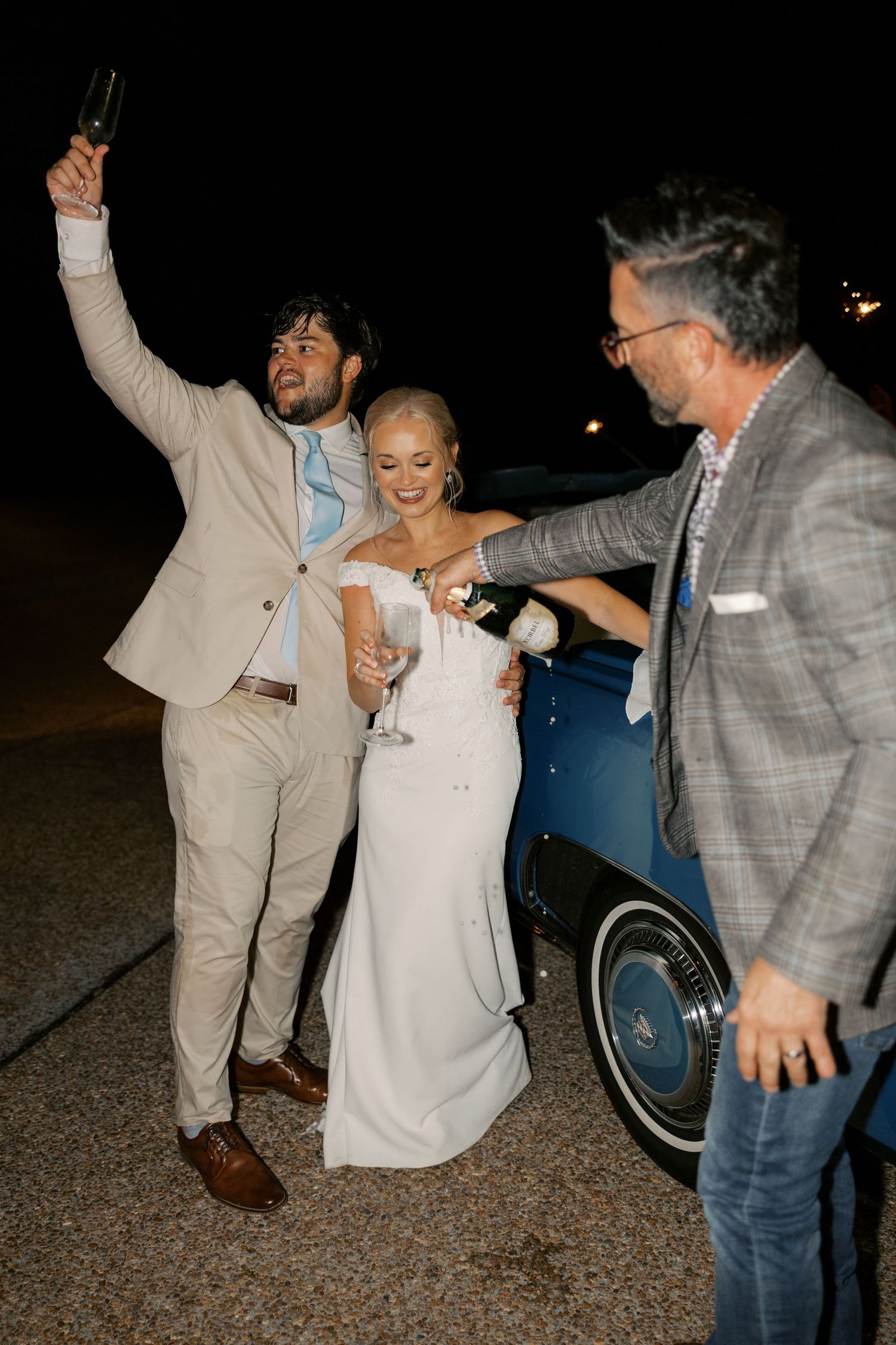 Wedding party celebrating with enthusiastic dance moves near a classic car at night.