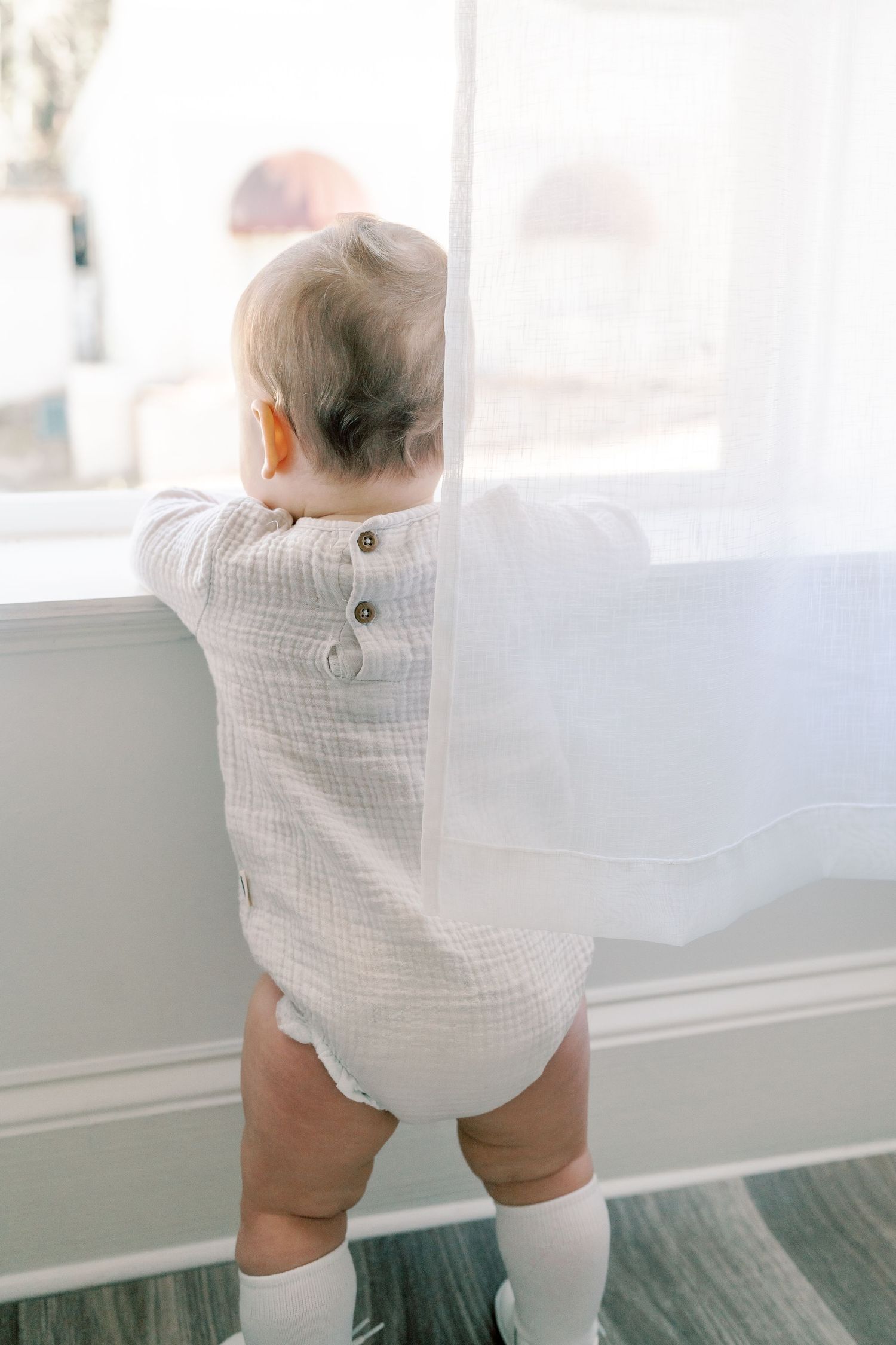 Toddler in white romper standing near window with flowing curtains.
