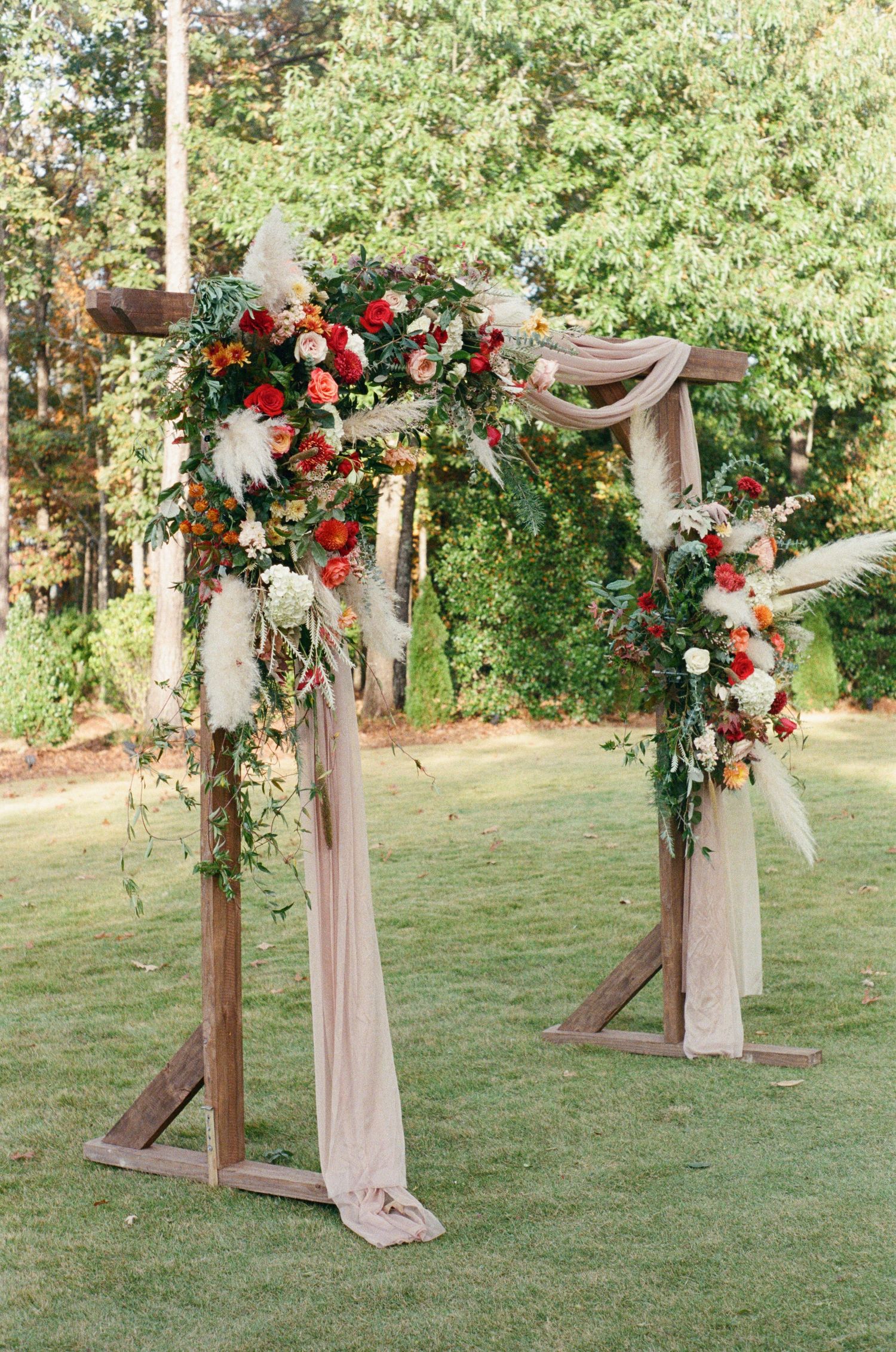 Wooden wedding arch decorated with blush fabric, red roses, and greenery on a grassy hillside.