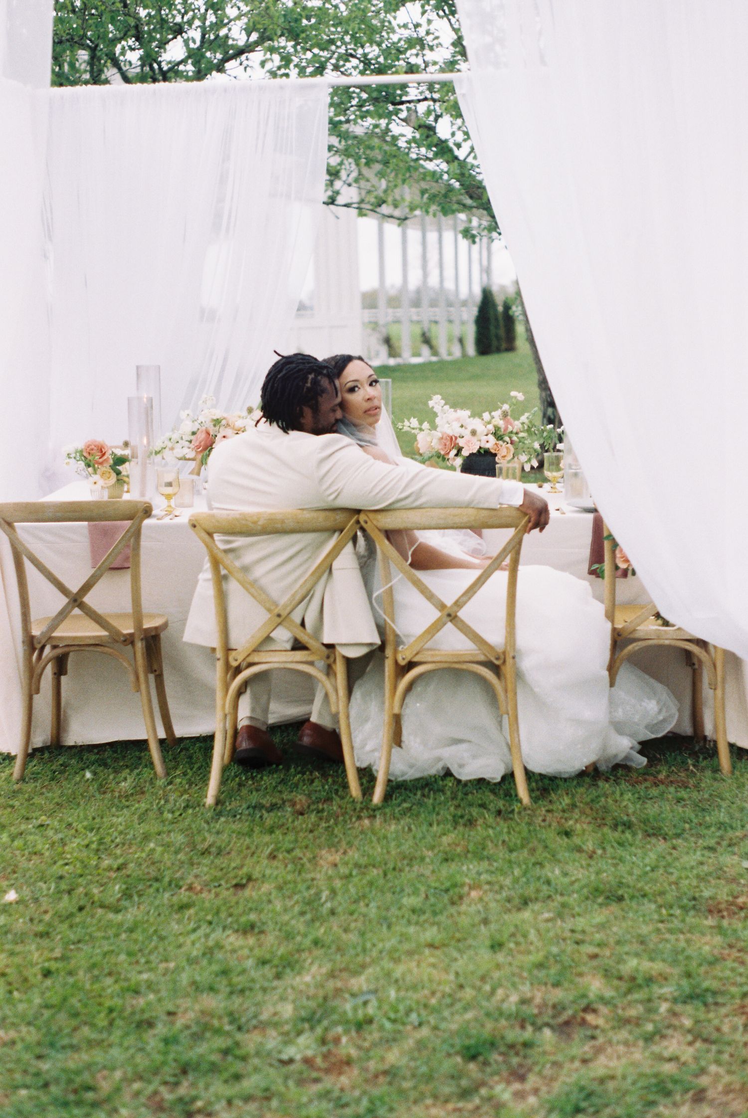 Outdoor wedding reception setup with white drapes and wooden chairs on grass.