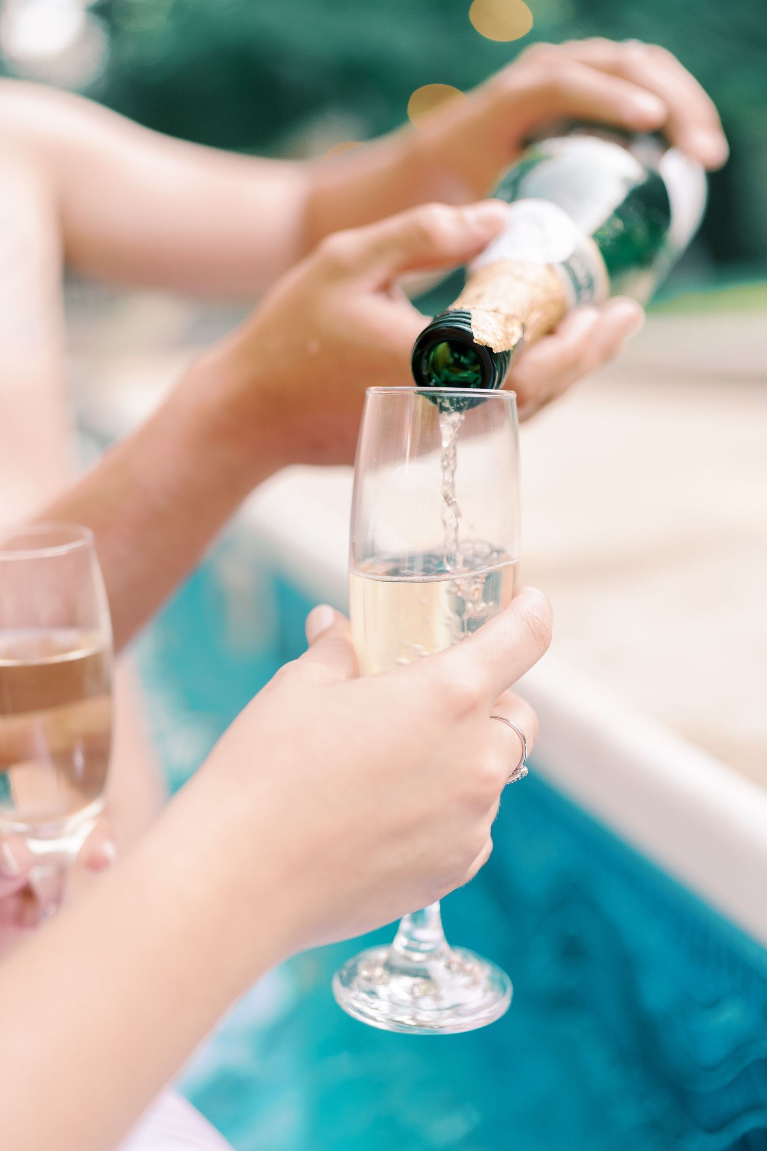 Close-up of hands pouring champagne into glasses by the poolside.