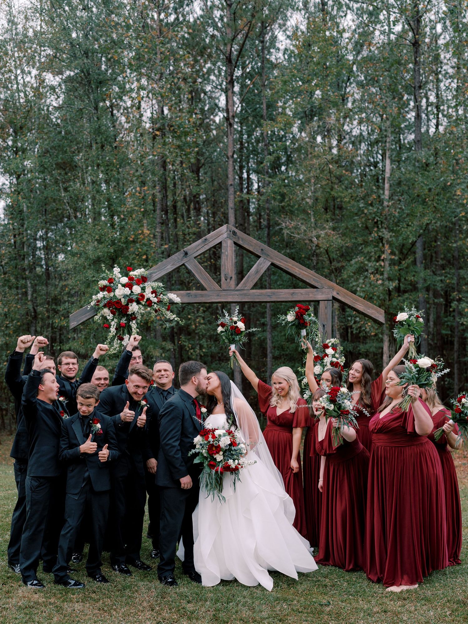 Wedding party in burgundy dresses and black suits celebrates outdoors with raised arms.