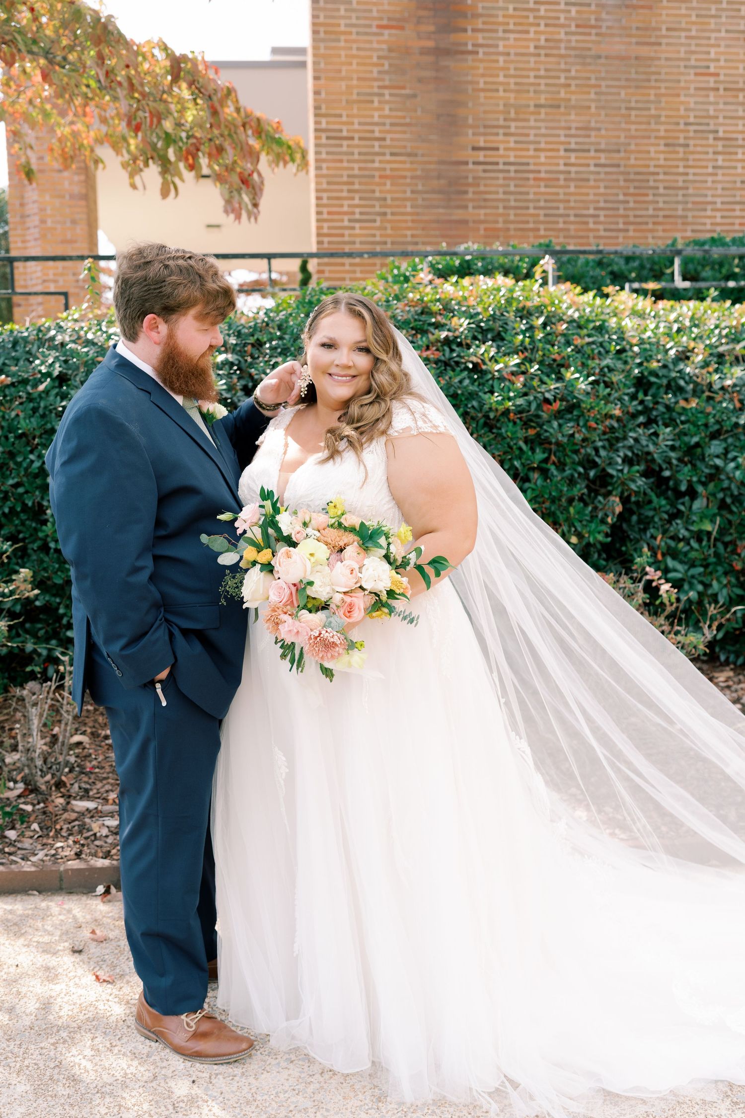 A couple poses for wedding photos with a long flowing veil against a brick backdrop.