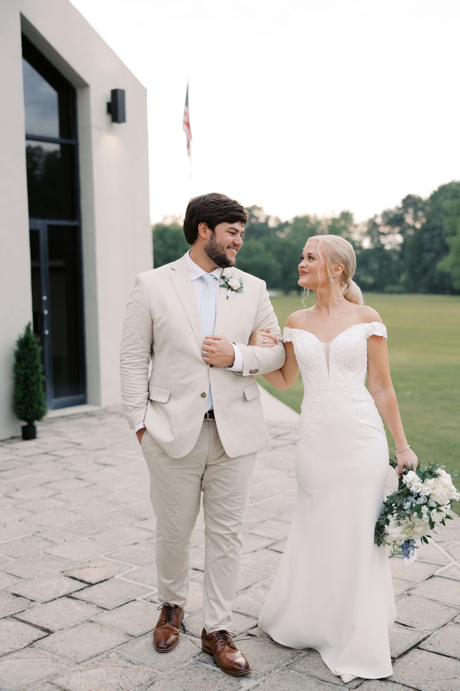 Wedding couple walking together on stone patio outside white building.