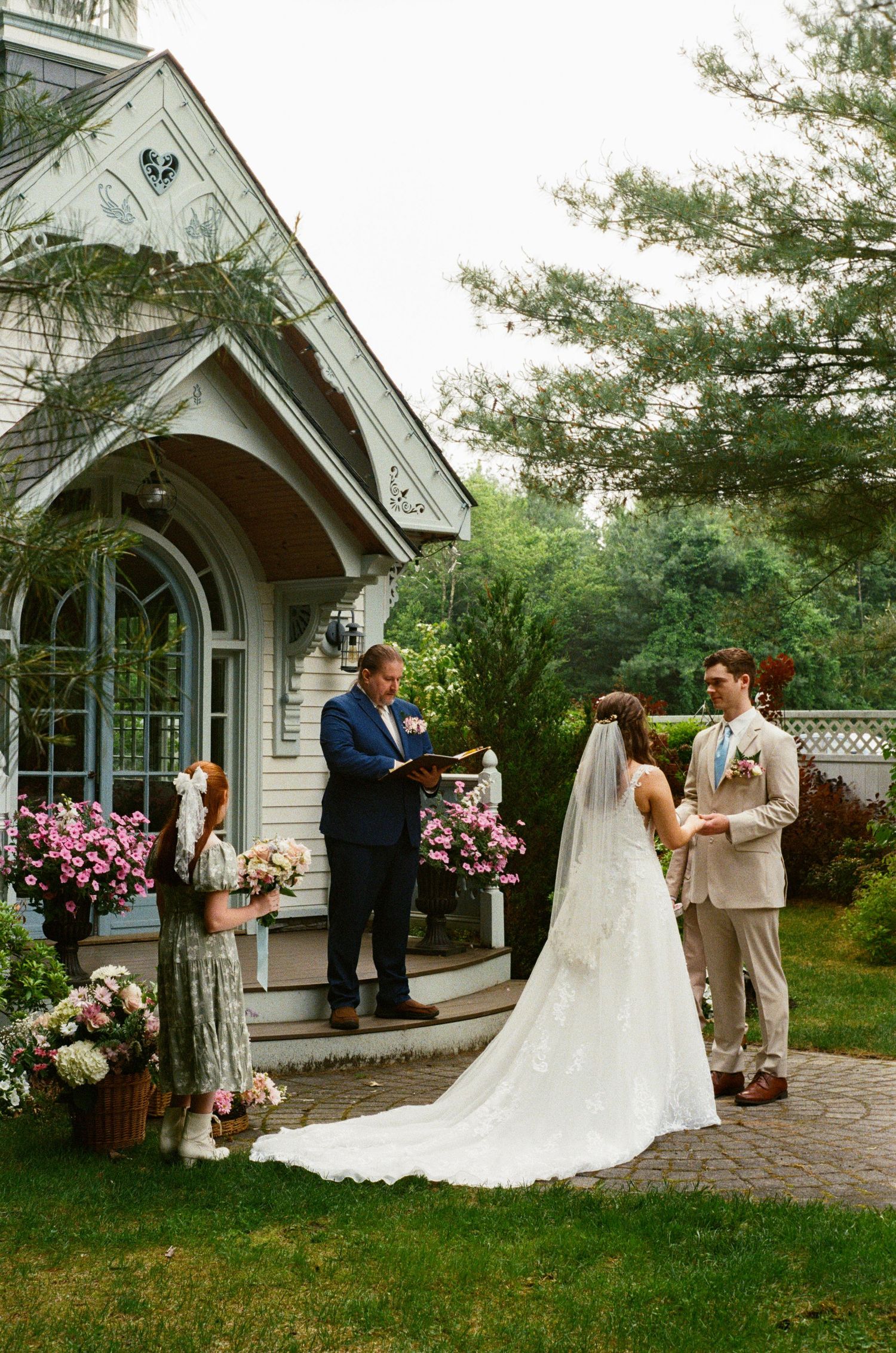 Wedding ceremony in progress outside white chapel with decorative pink flower arrangements.