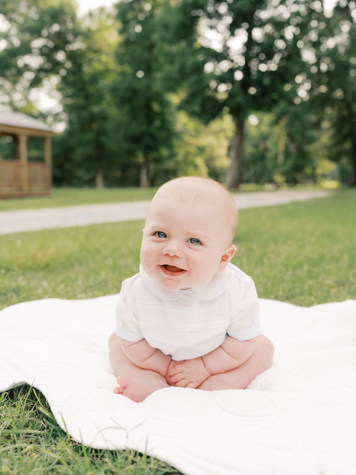 Adorable baby in white clothing smiling while laying on blanket in park setting.