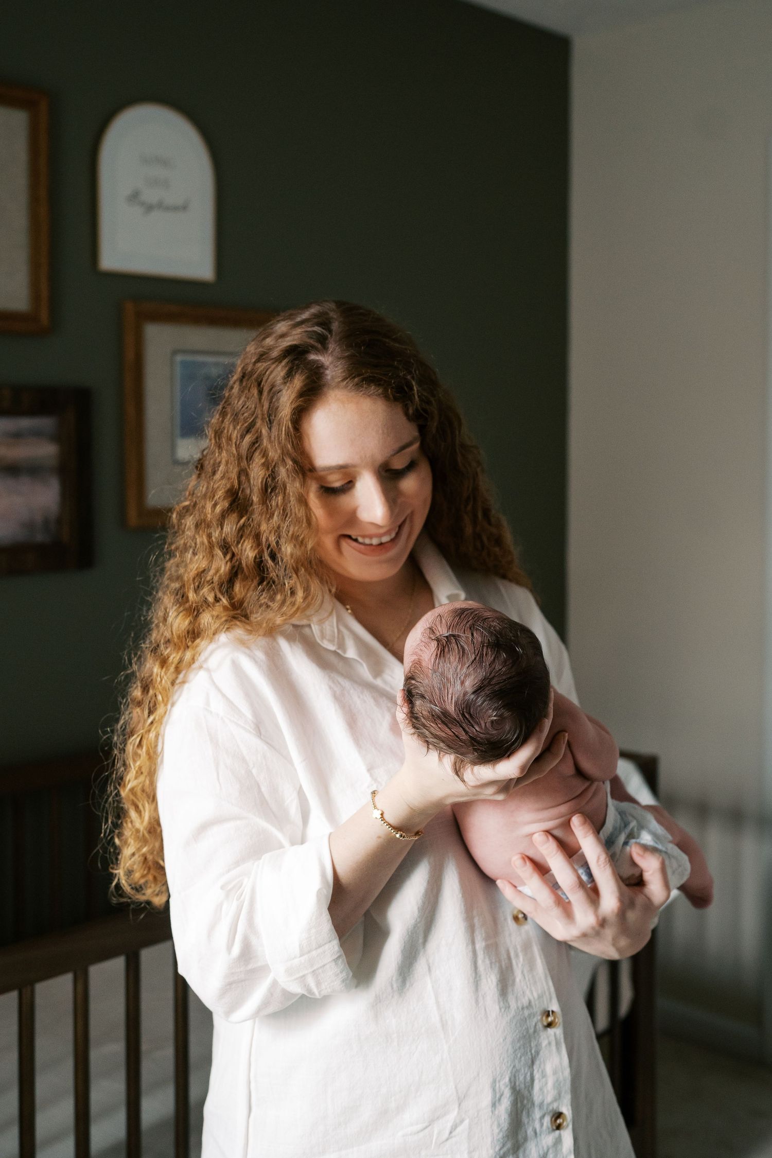 Woman in white holding and kissing newborn baby in front of dark green wall with natural window light.