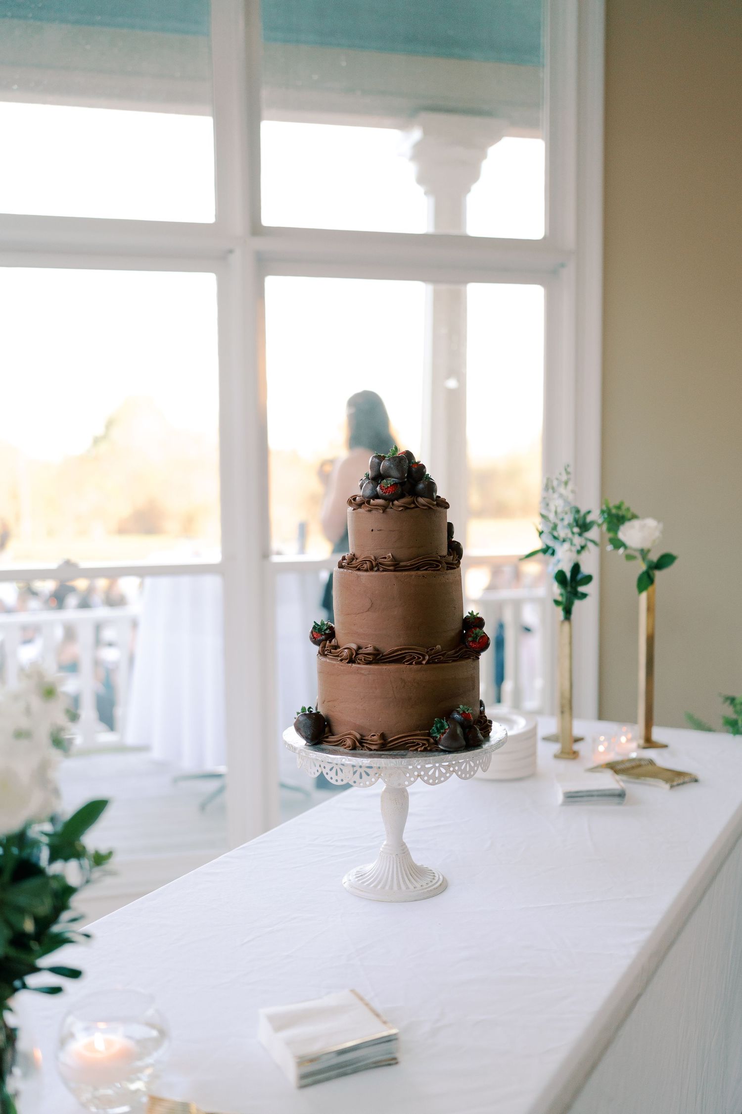 Chocolate wedding cake displayed on white pedestal stand in bright sunlit reception room.