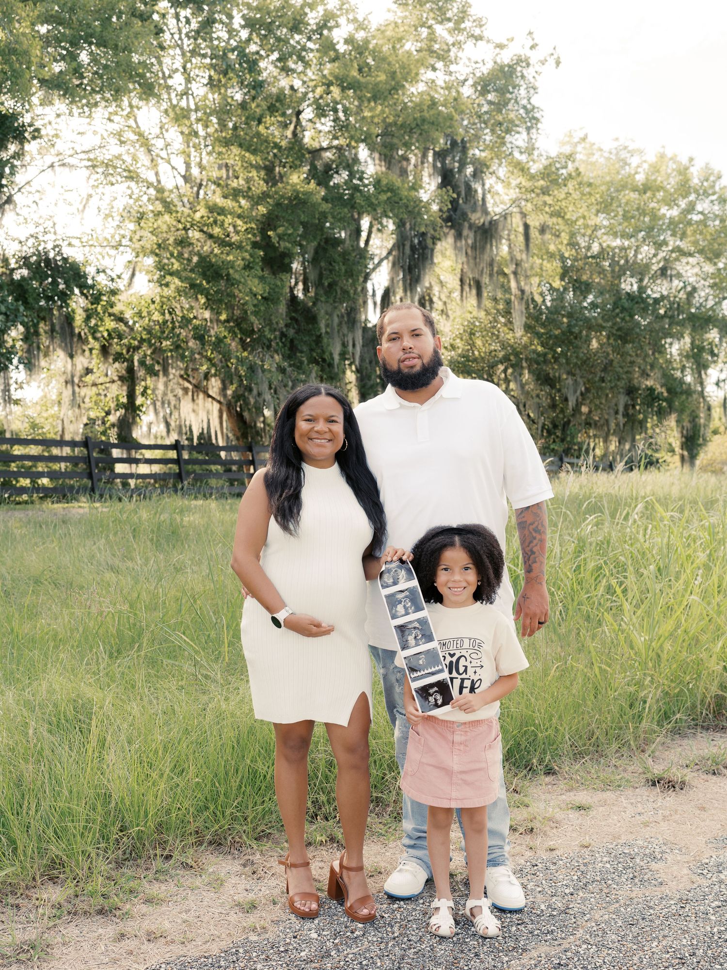 A family holds ultrasound photos while standing on a dirt path in a grassy field.