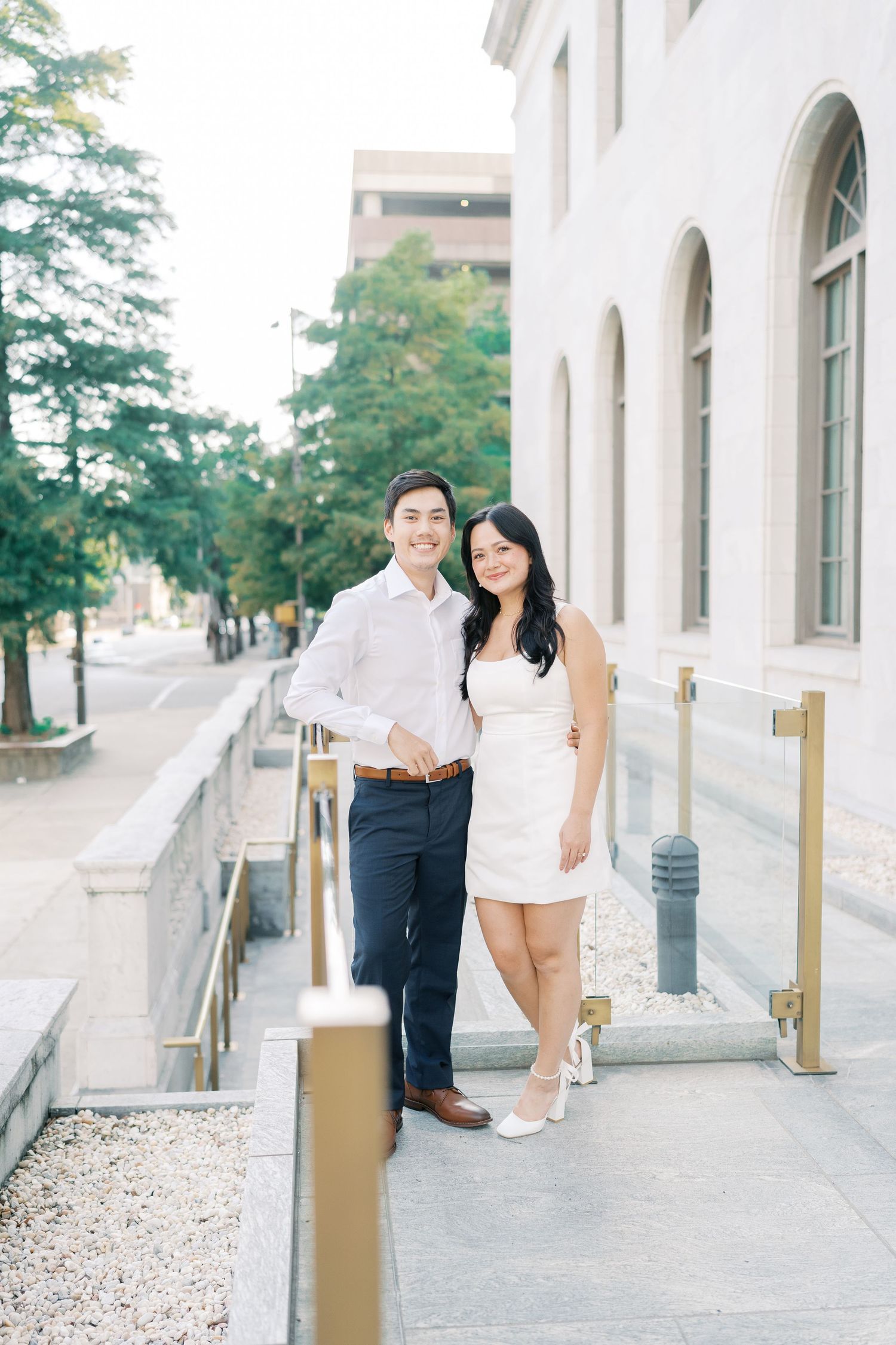 A couple poses casually along a building's exterior walkway with columns and trees in background.