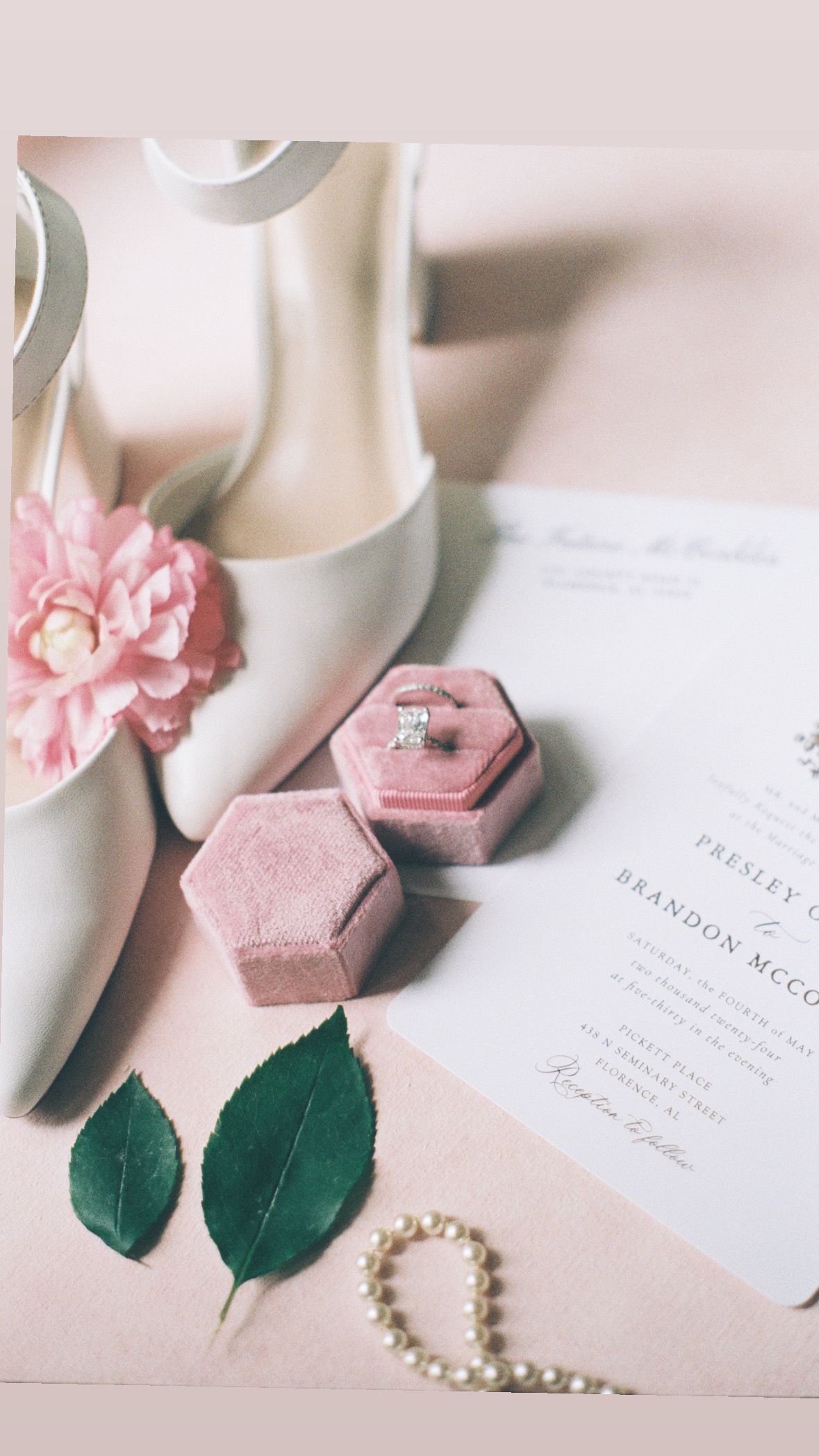Wedding details featuring white shoes, pink floral accents, and invitation on light background.