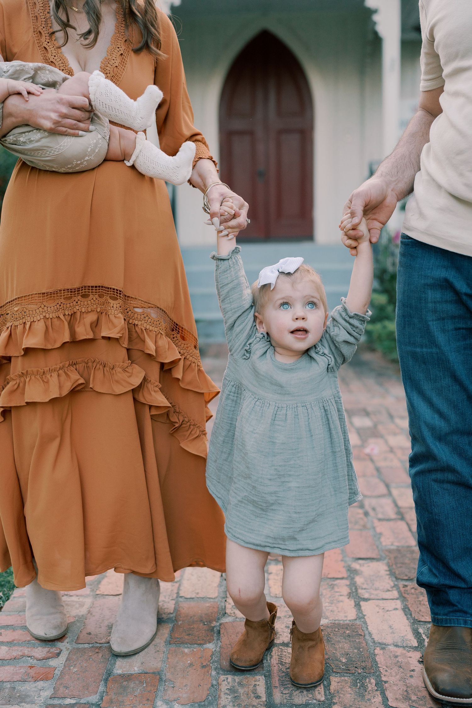 A toddler in a gray dress takes first steps between parents on brick pavement.