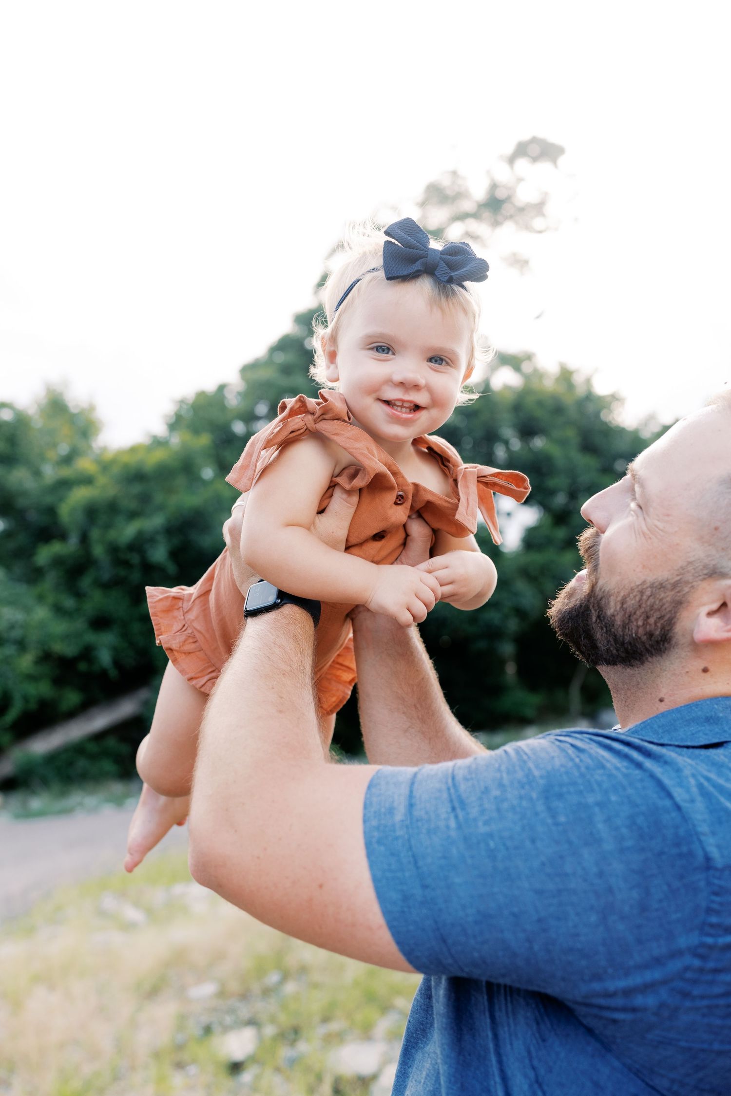 A parent lifts a small child wearing orange clothing against a natural outdoor backdrop.