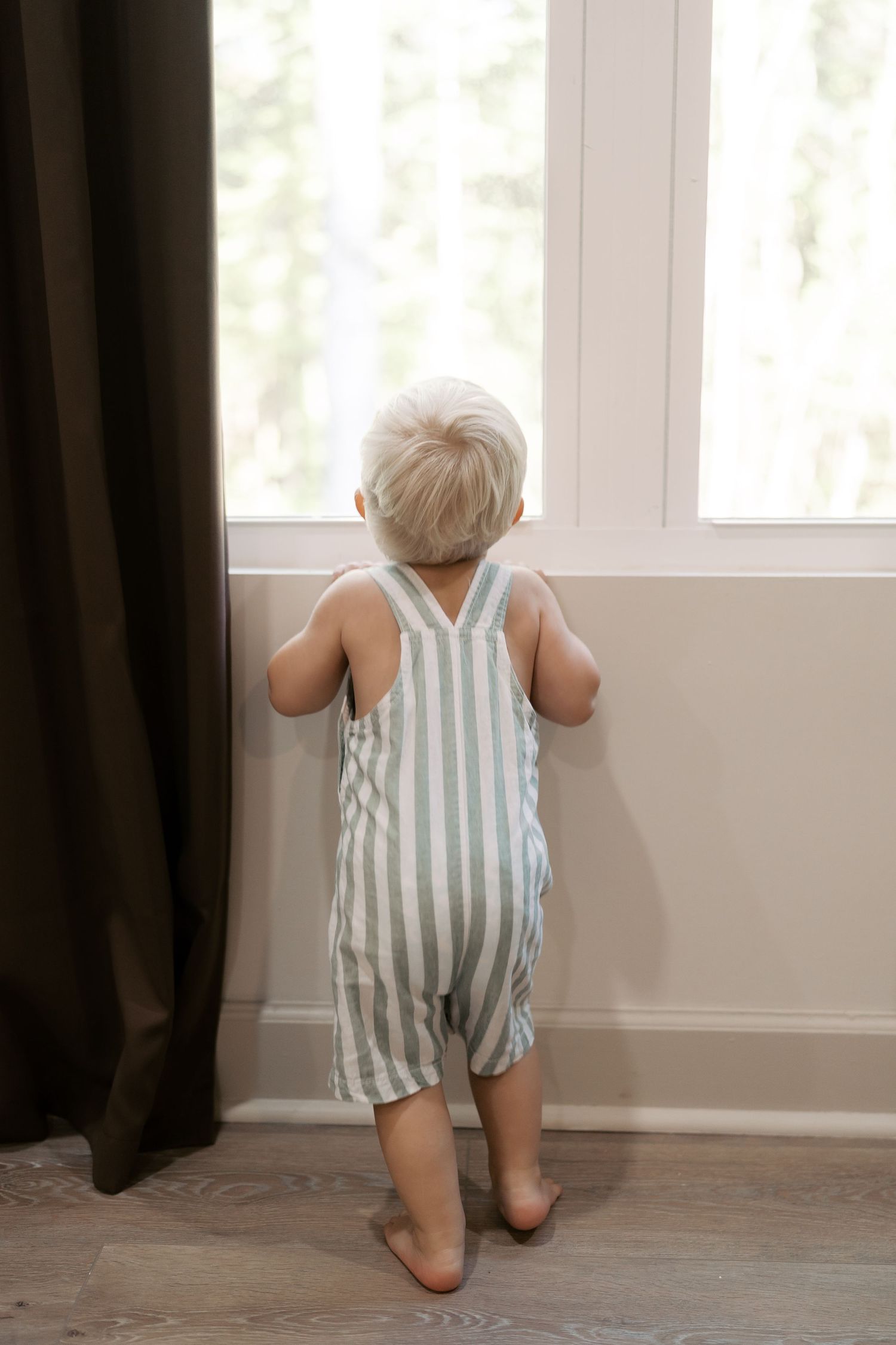 Small child in striped romper looking out window with dark curtains.
