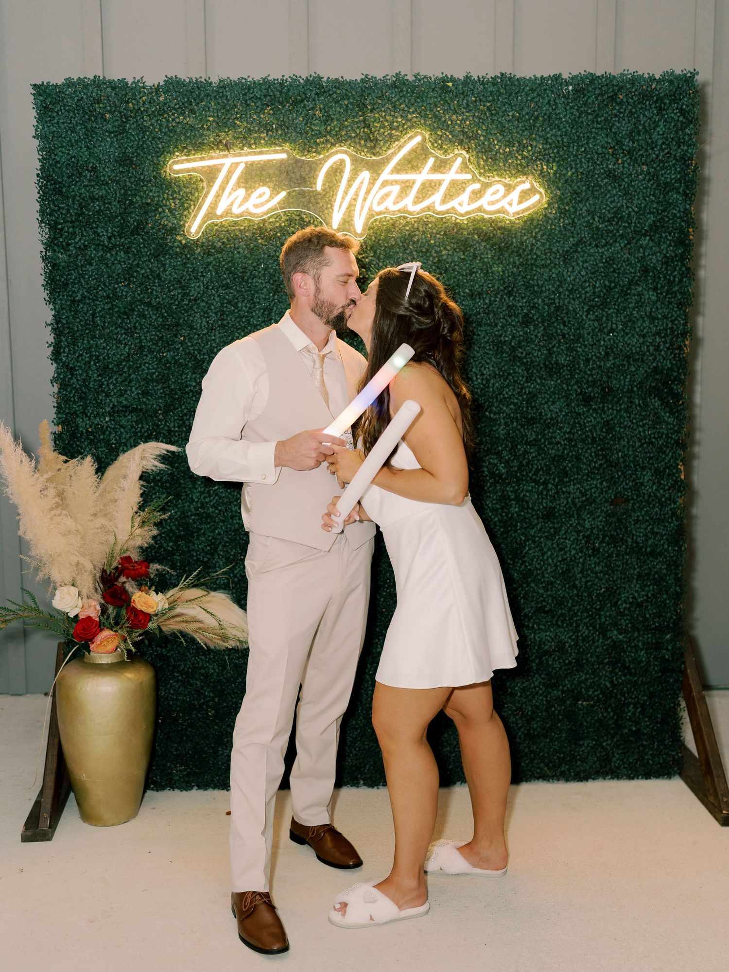 Couple poses in front of green hedge wall with neon 'The Mattress' sign at wedding reception.
