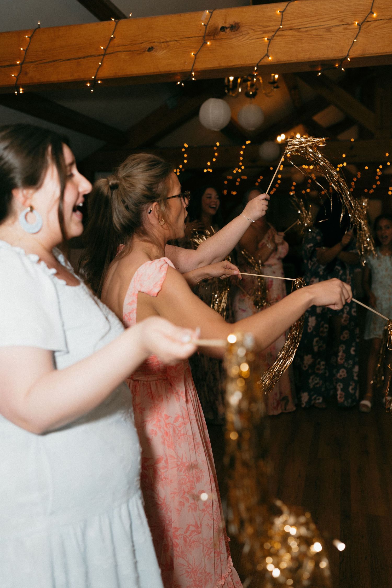 Wedding guests dance together in a rustic venue decorated with twinkling lights and wooden beams.