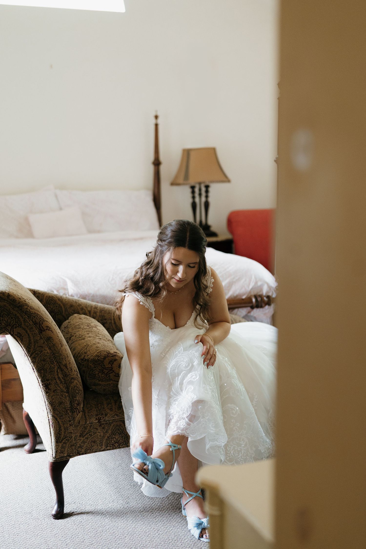 Someone in a white wedding dress adjusting their blue garter while sitting on a brown chair.