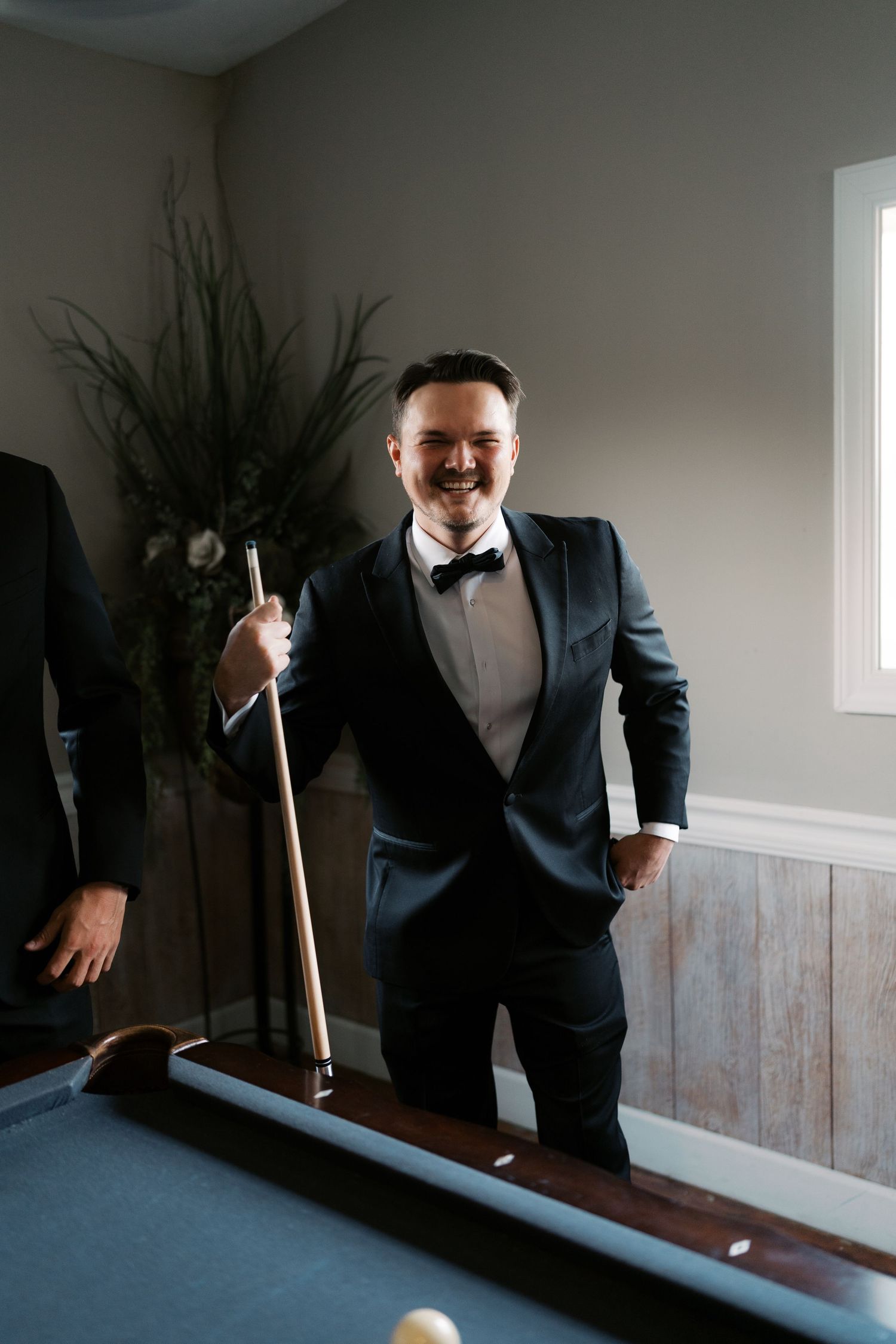 Groom in black tuxedo holding pool cue and smiling in game room.