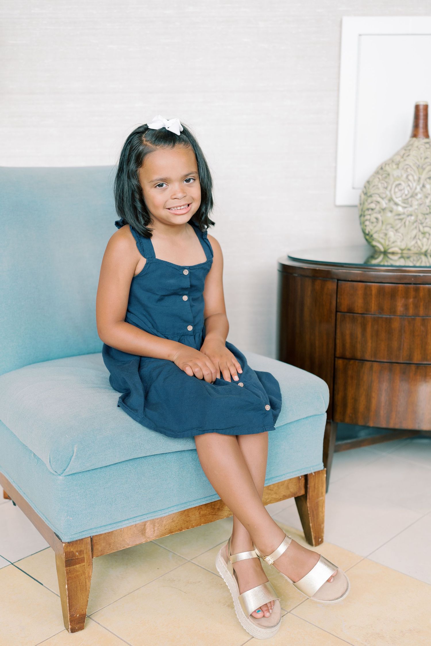 Young child sitting on a light blue bench wearing a navy dress and white sandals.