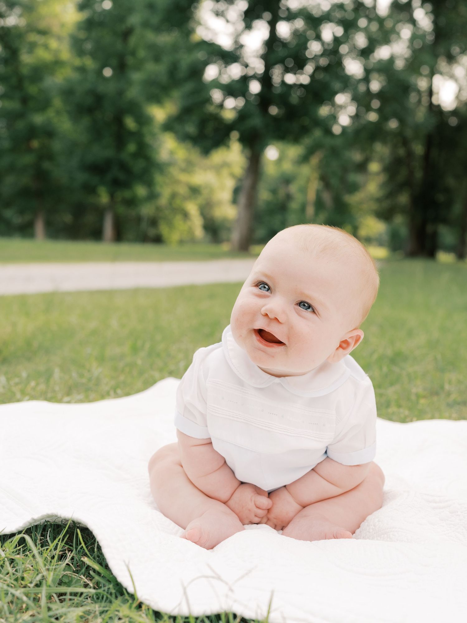 Baby in white outfit lying on blanket outdoors with bright eyes and sweet expression.