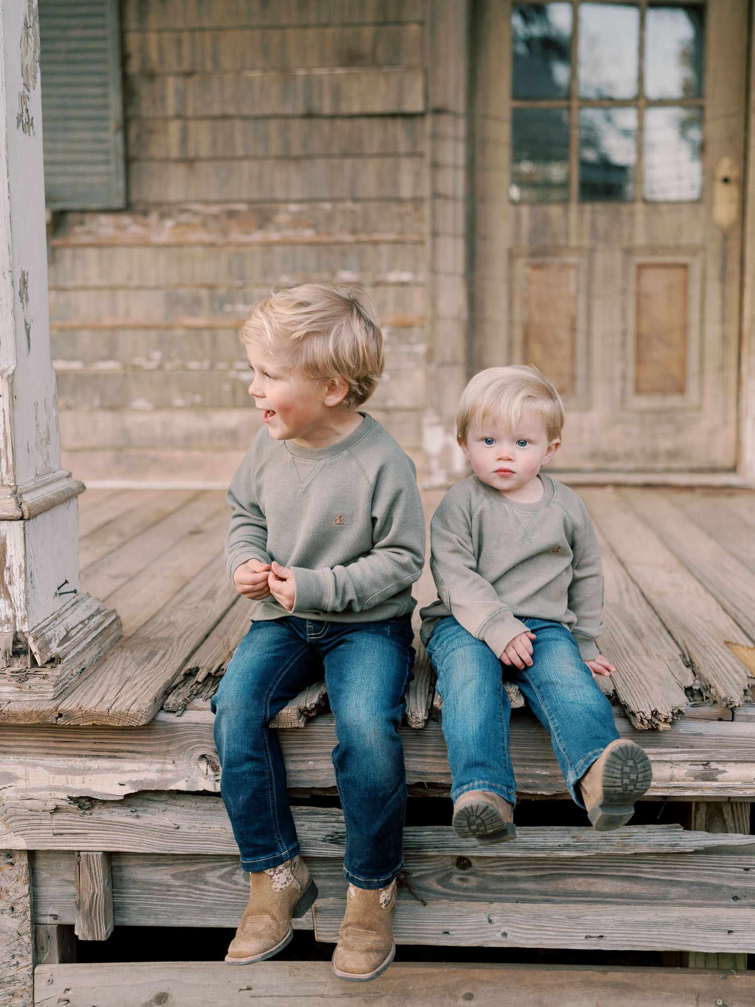 Two children in matching gray sweaters sit on a rustic wooden porch.