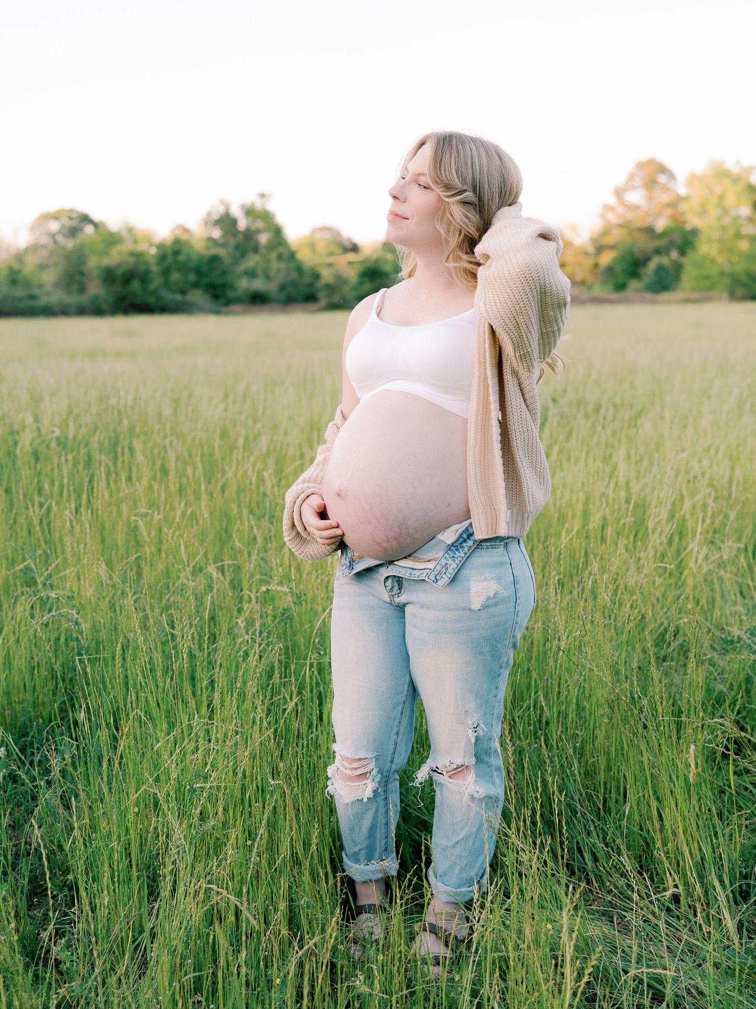 Maternity photos showing a pregnant woman in ripped jeans and white top standing in a sunlit meadow of tall grass.