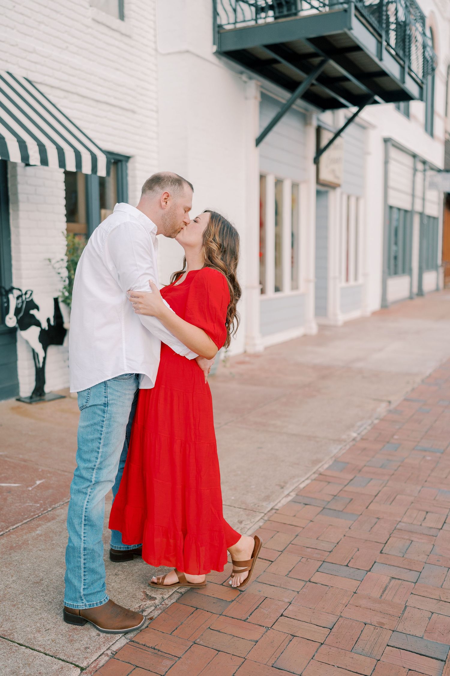 A romantic couple shares a tender moment on a brick-lined street in front of white buildings.