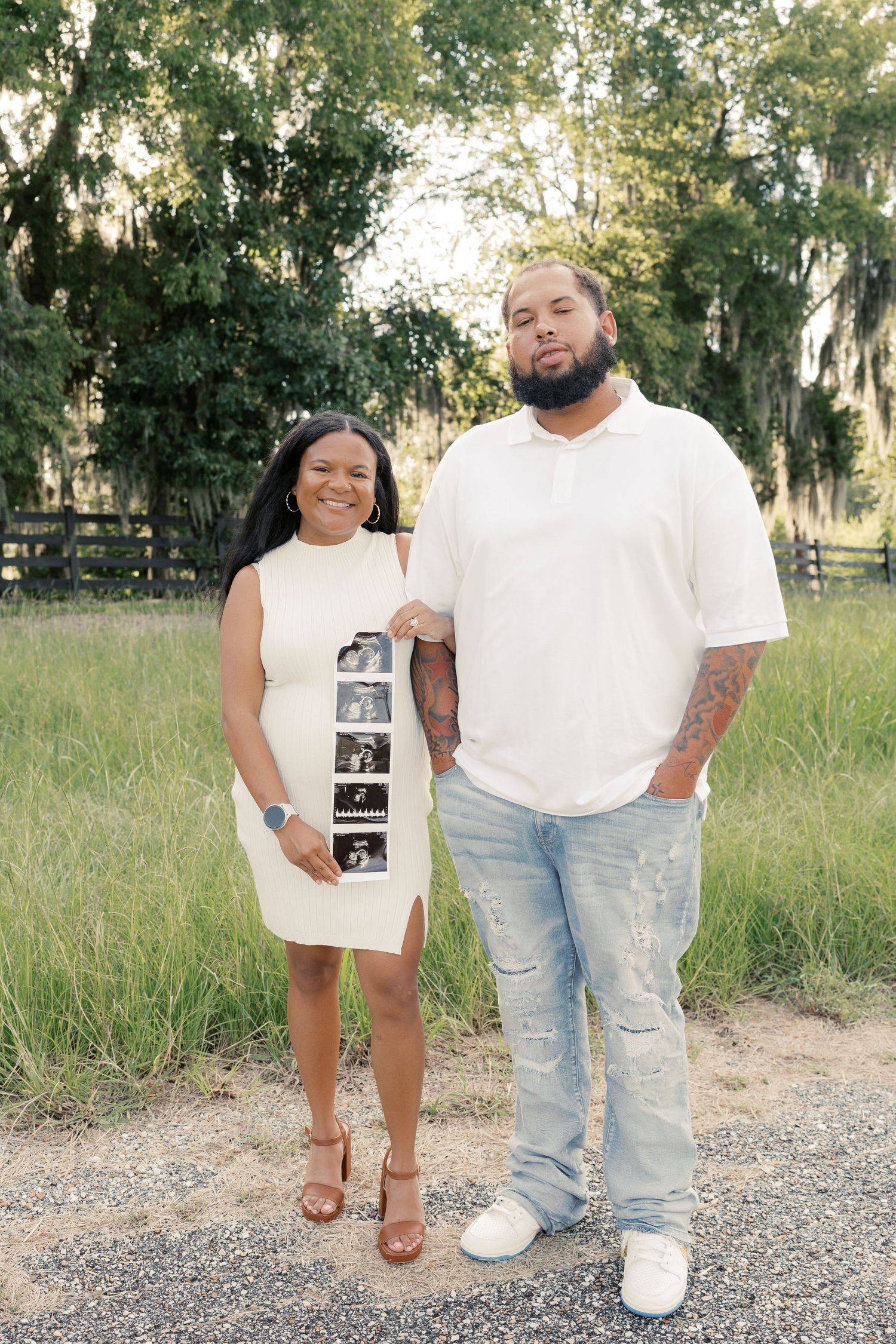 A family poses together holding ultrasound photos on a gravel path during sunset.