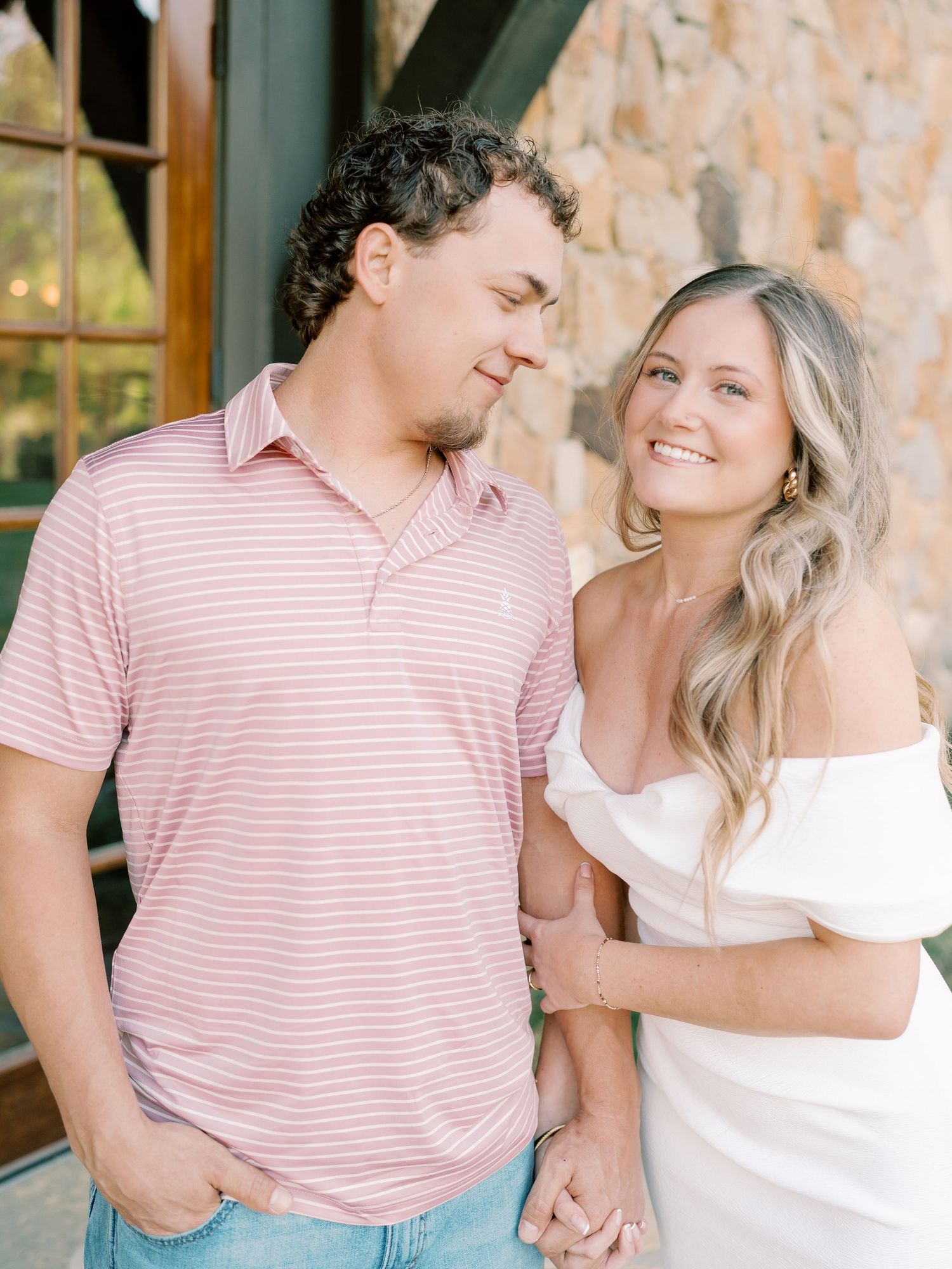 Couple shares romantic moments in front of a rustic stone wall wearing pink and white outfits.