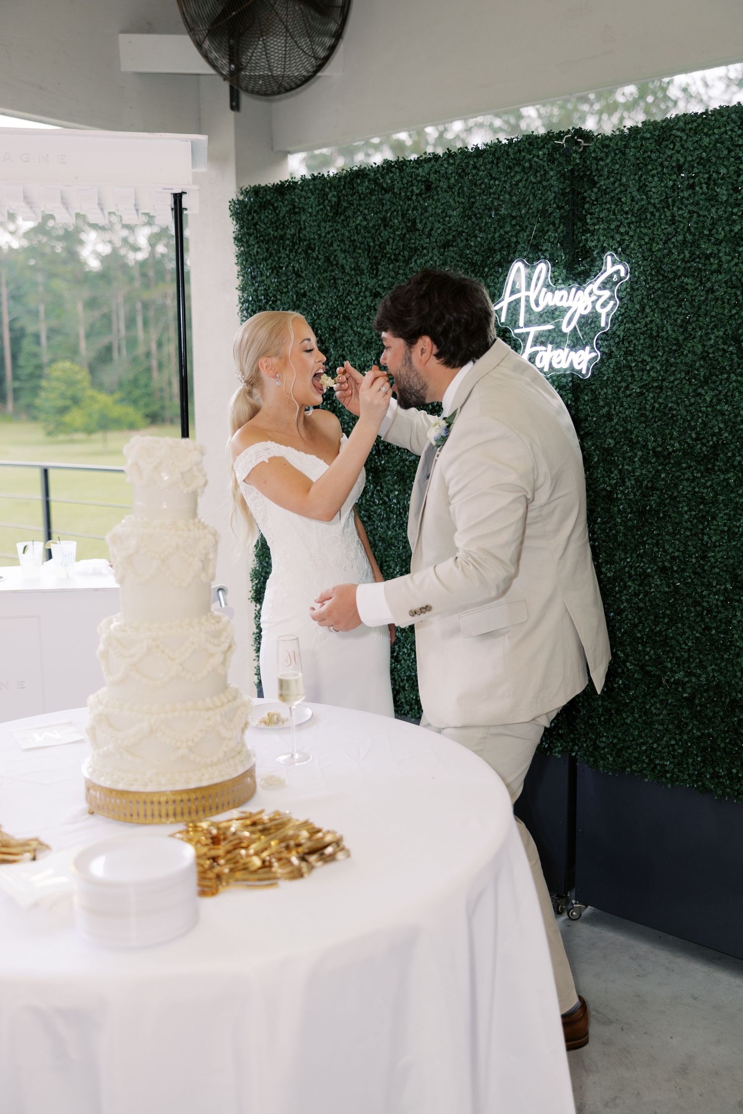 Wedding cake cutting ceremony with white cake and green hedge wall backdrop.