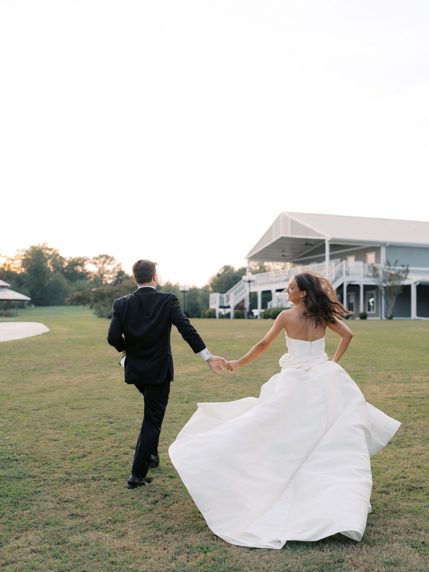 Wedding couple running playfully across grassy field with flowing white dress.