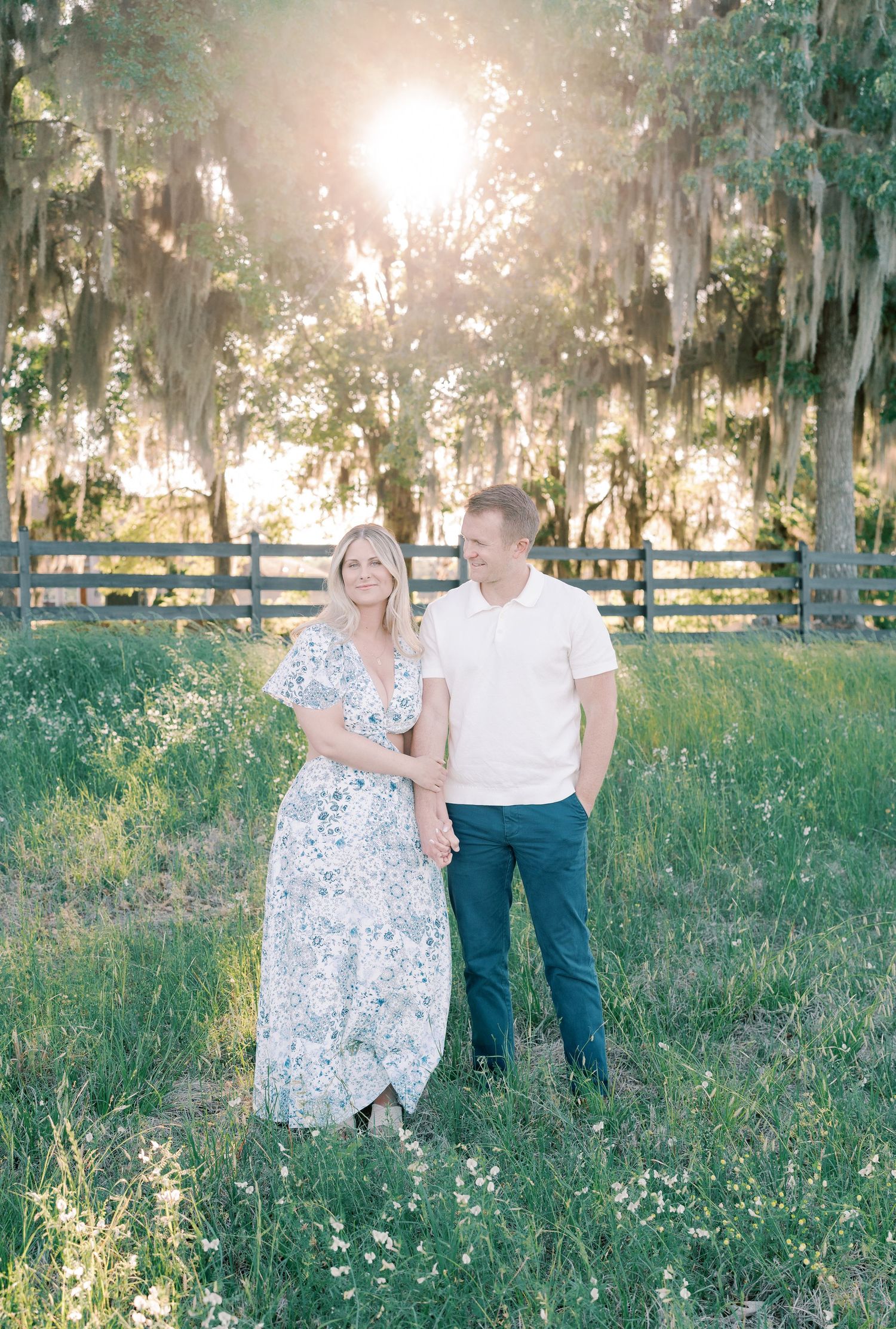 A couple poses together in a sunlit field with tall grass and Spanish moss draped trees.