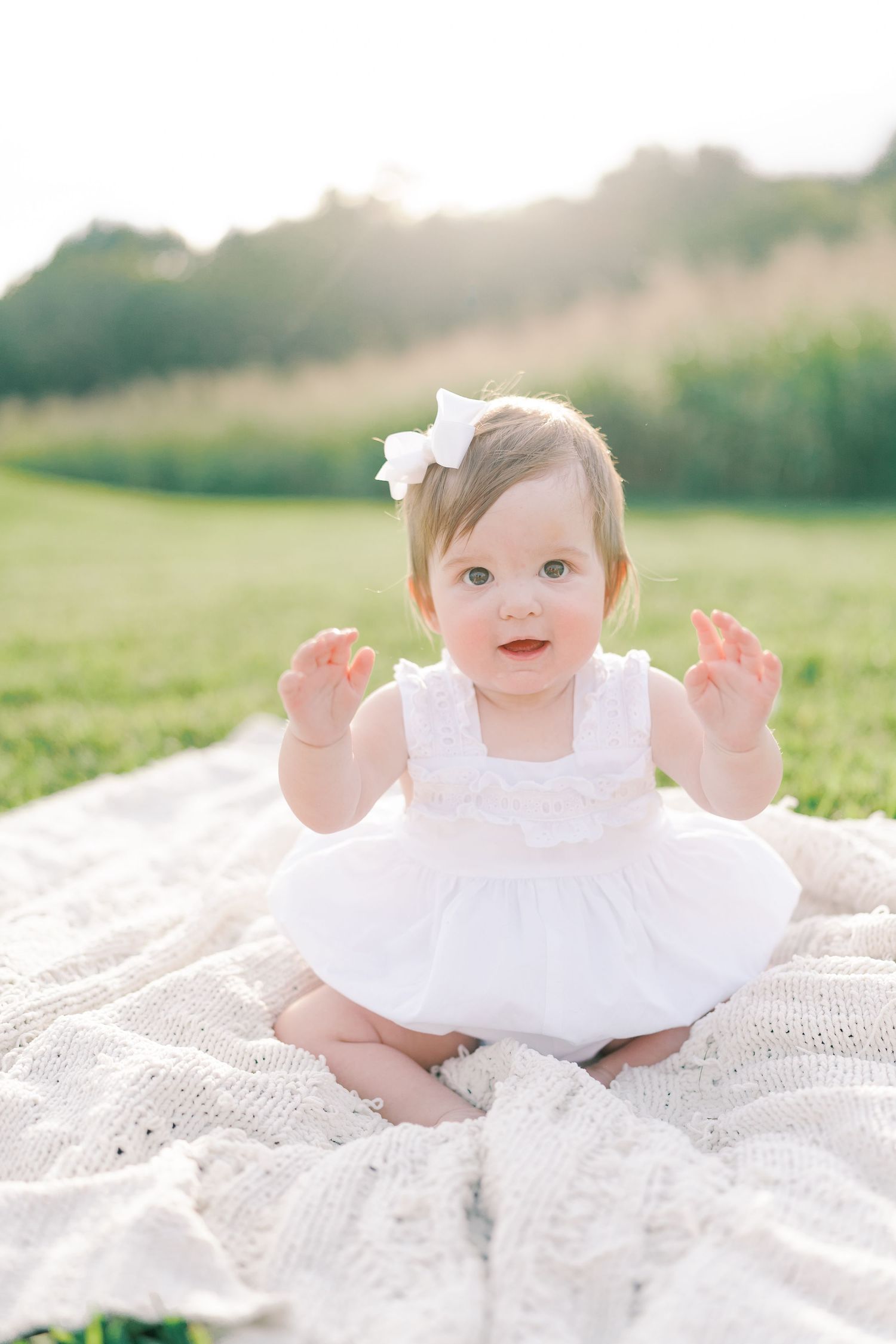 Baby in white dress sitting on lace blanket outdoors during golden hour photo session.