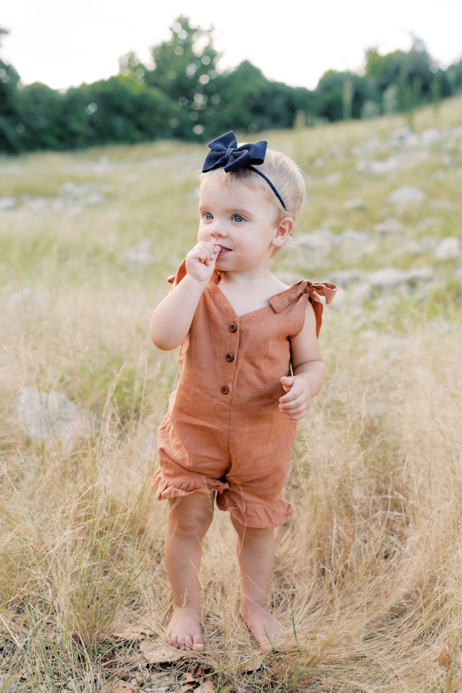 Toddler in brown romper and navy bow standing in golden field.