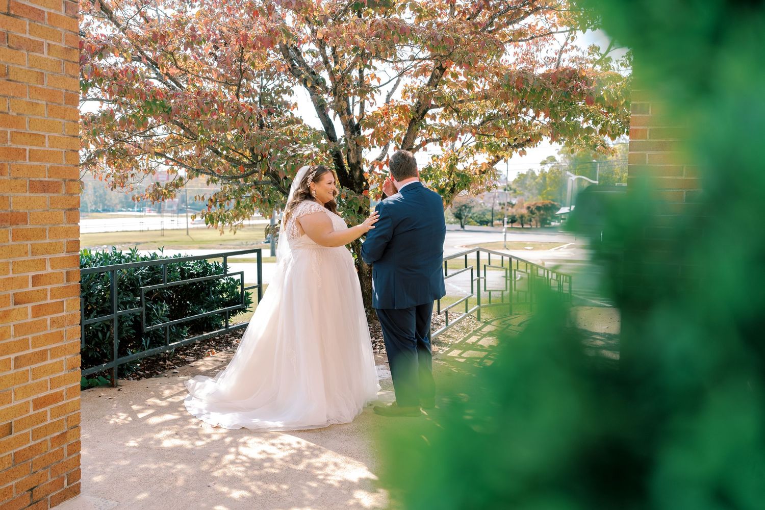 A couple shares a romantic moment on their wedding day against a backdrop of autumn trees and brick columns.