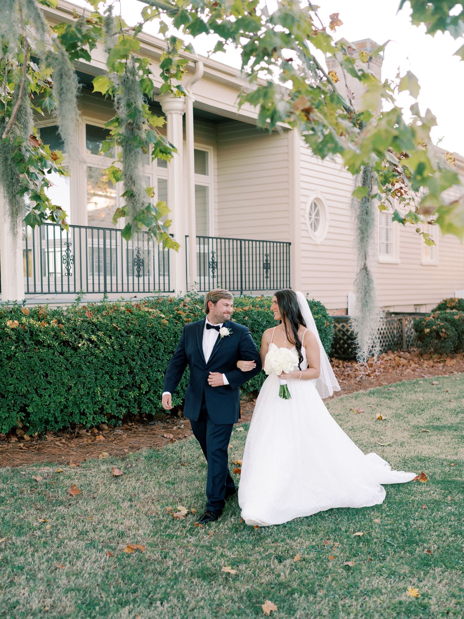 Wedding couple walking together on a lawn in front of a building with green shutters.