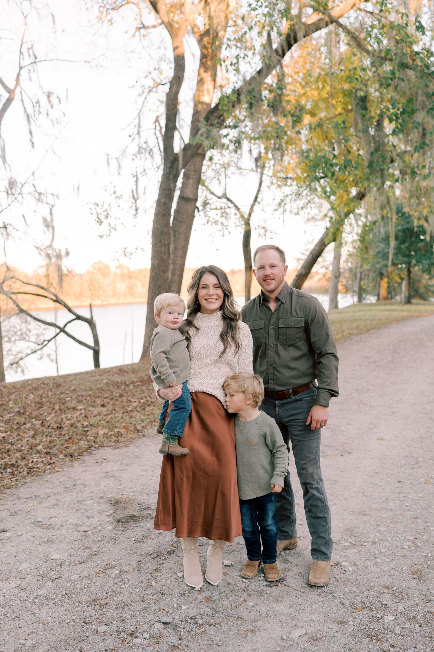 A family poses together on a dirt path surrounded by autumn trees near a lake.